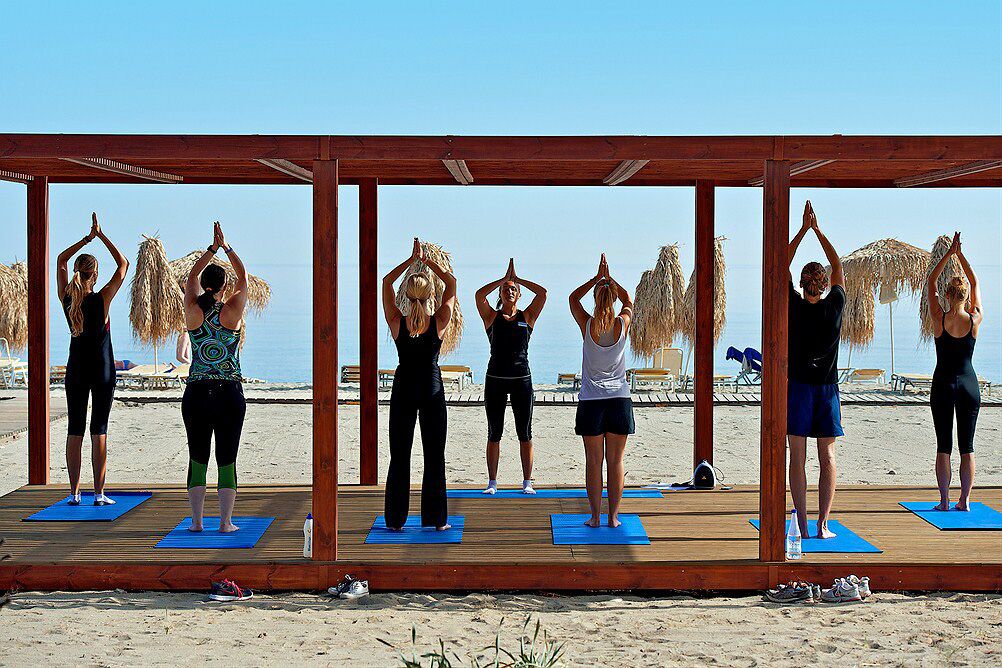 A group of people performing yoga on mats under a wooden pergola on a beach.