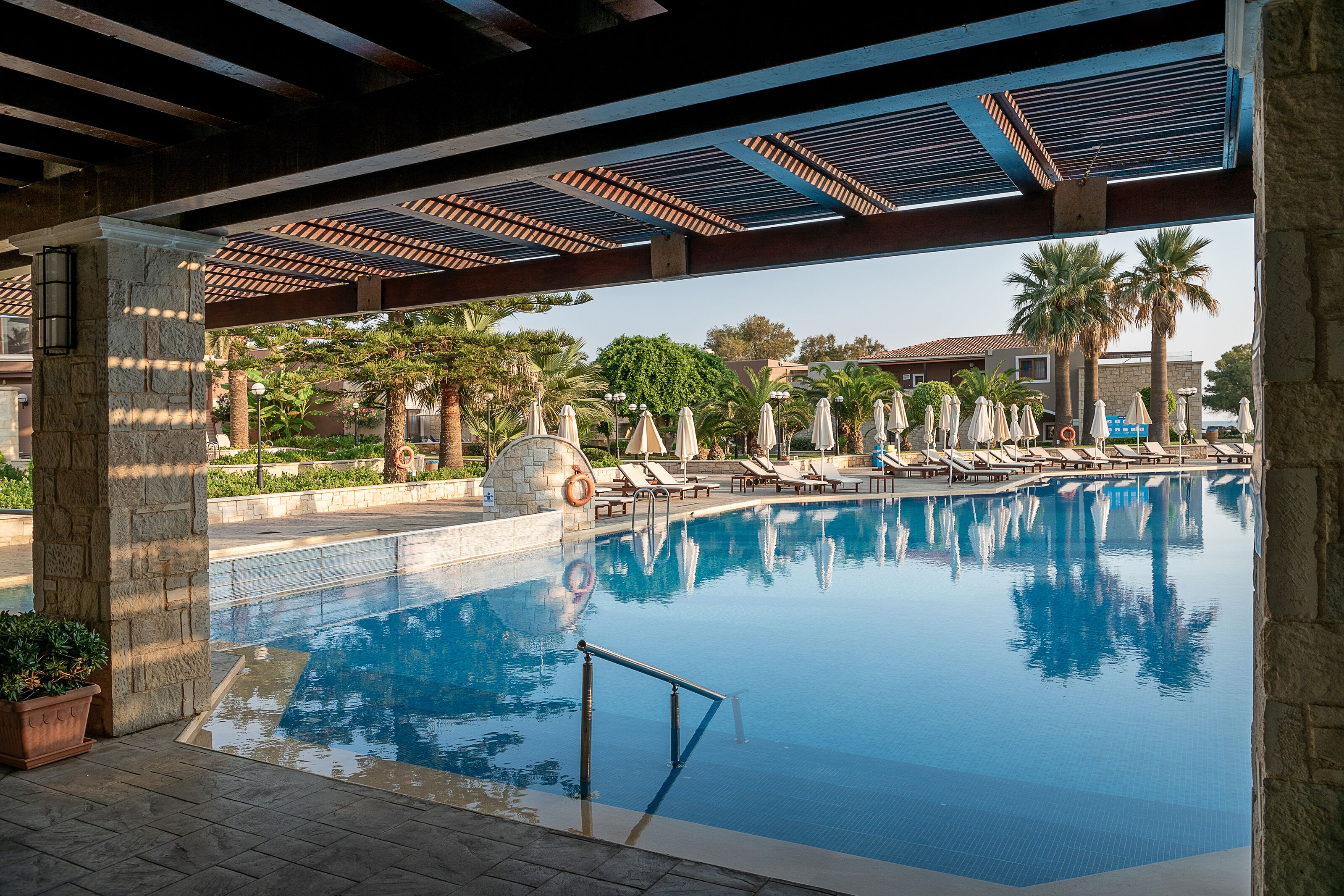 Piscina con sillas de descanso y sombrillas, rodeada de palmeras y una zona cubierta con techo de tejas.