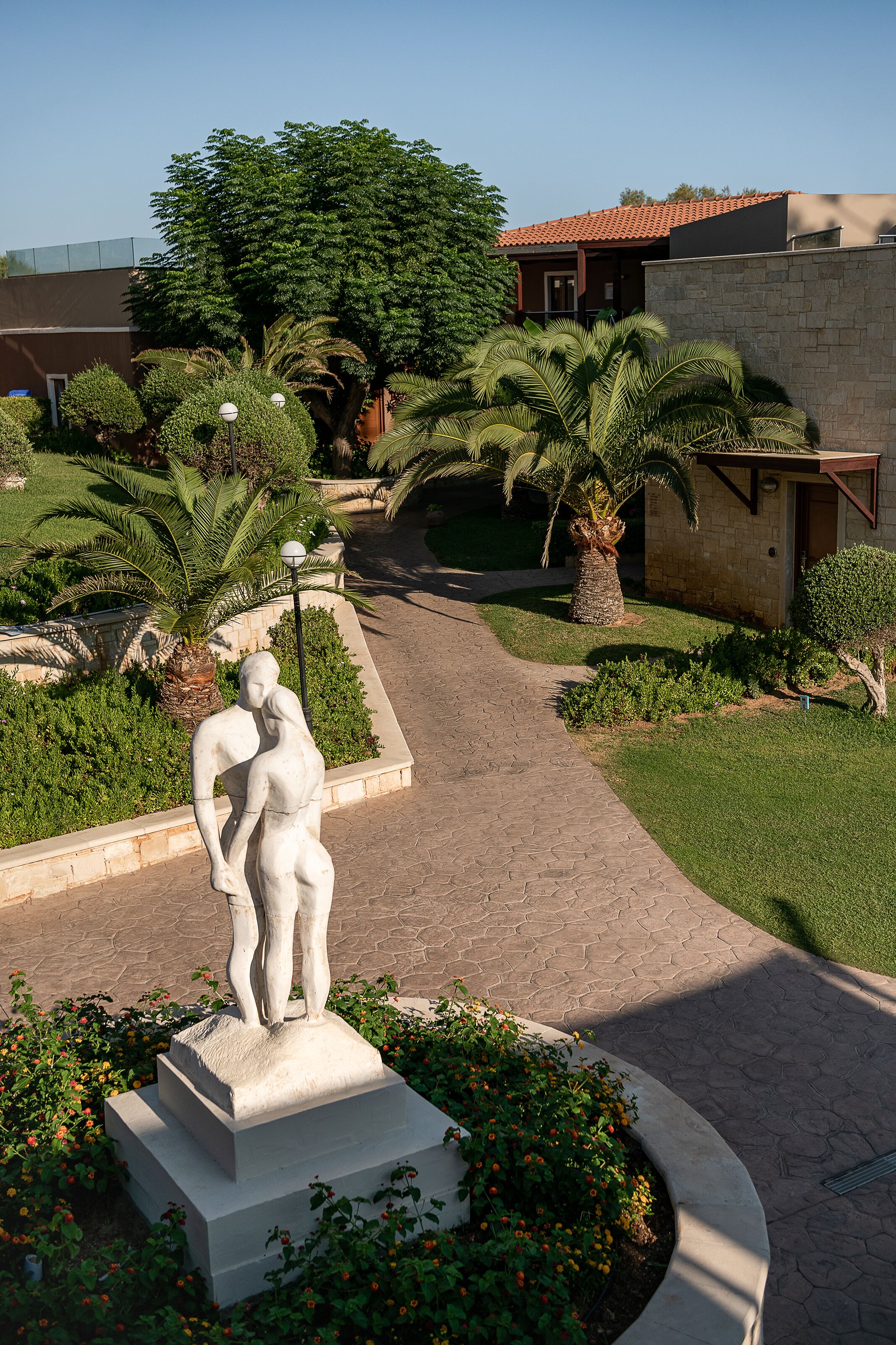 Una estatua en un jardín ajardinado con un sendero pavimentado, palmeras y un edificio con techo de tejas rojas.
