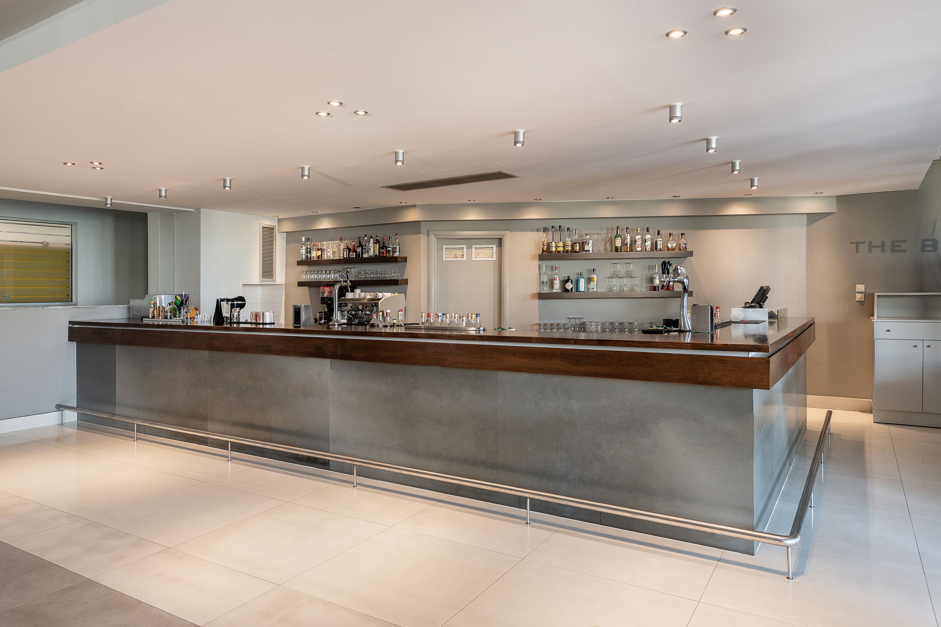 A modern bar area with a long counter, shelves with bottles, and a clean, tiled floor.