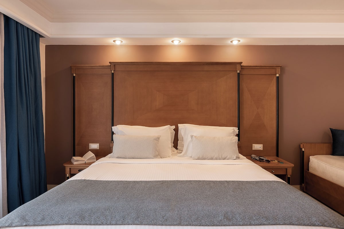 A hotel room with a large bed, two bedside tables, a wooden headboard, and a nightstand with a phone and book.