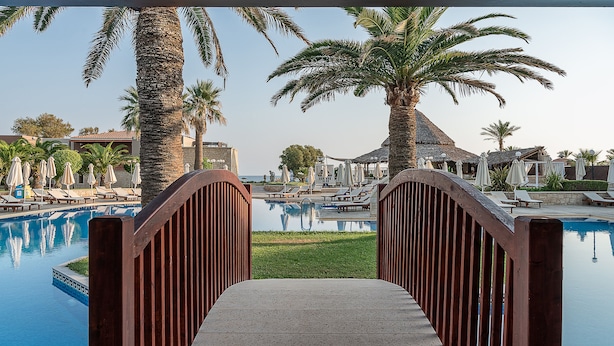A pool area with lounge chairs and umbrellas, a wooden bridge, and palm trees. - Caldera Creta Paradise (Platanias)