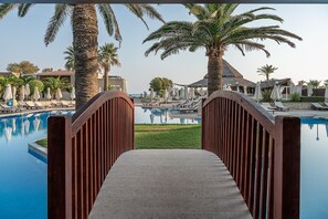 A pool area with lounge chairs and umbrellas, a wooden bridge, and palm trees. - Caldera Creta Paradise (Platanias)