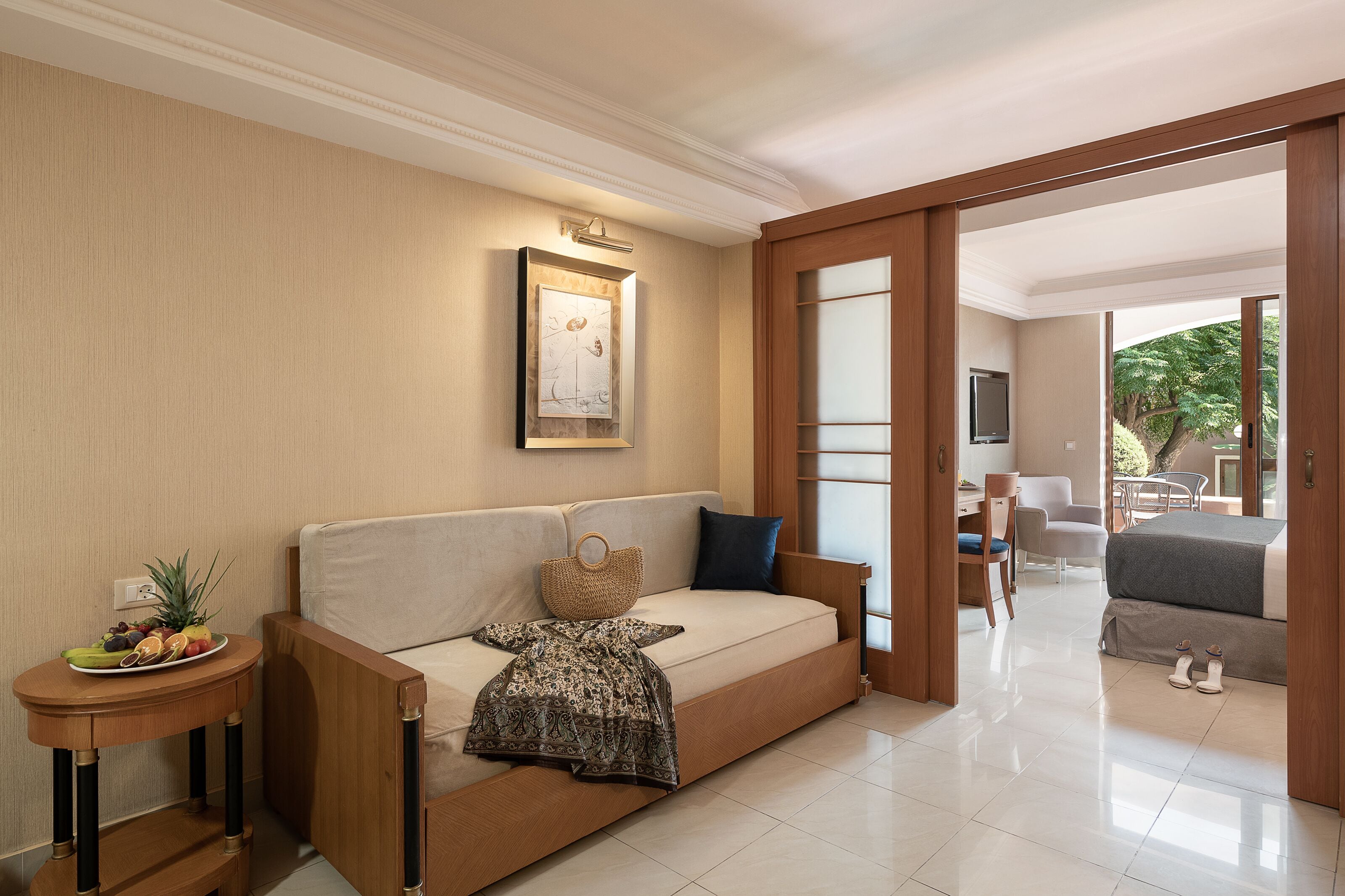 A modern hotel room with a sofa, a small side table with fruits, a framed picture, and a view of the outdoor area.