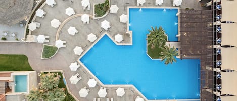 Vista aérea de um resort com uma piscina central, cercada por guarda-sóis brancos e espreguiçadeiras, e uma área separada com piscina e espreguiçadeiras.