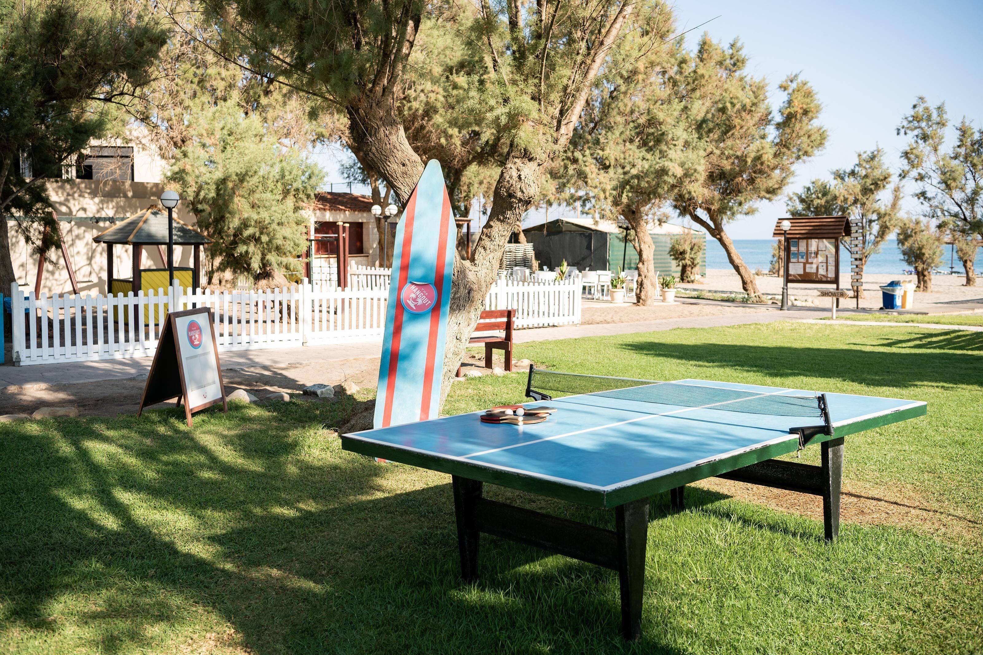 Una mesa de ping pong en un parque con una tabla de surf apoyada en un árbol.