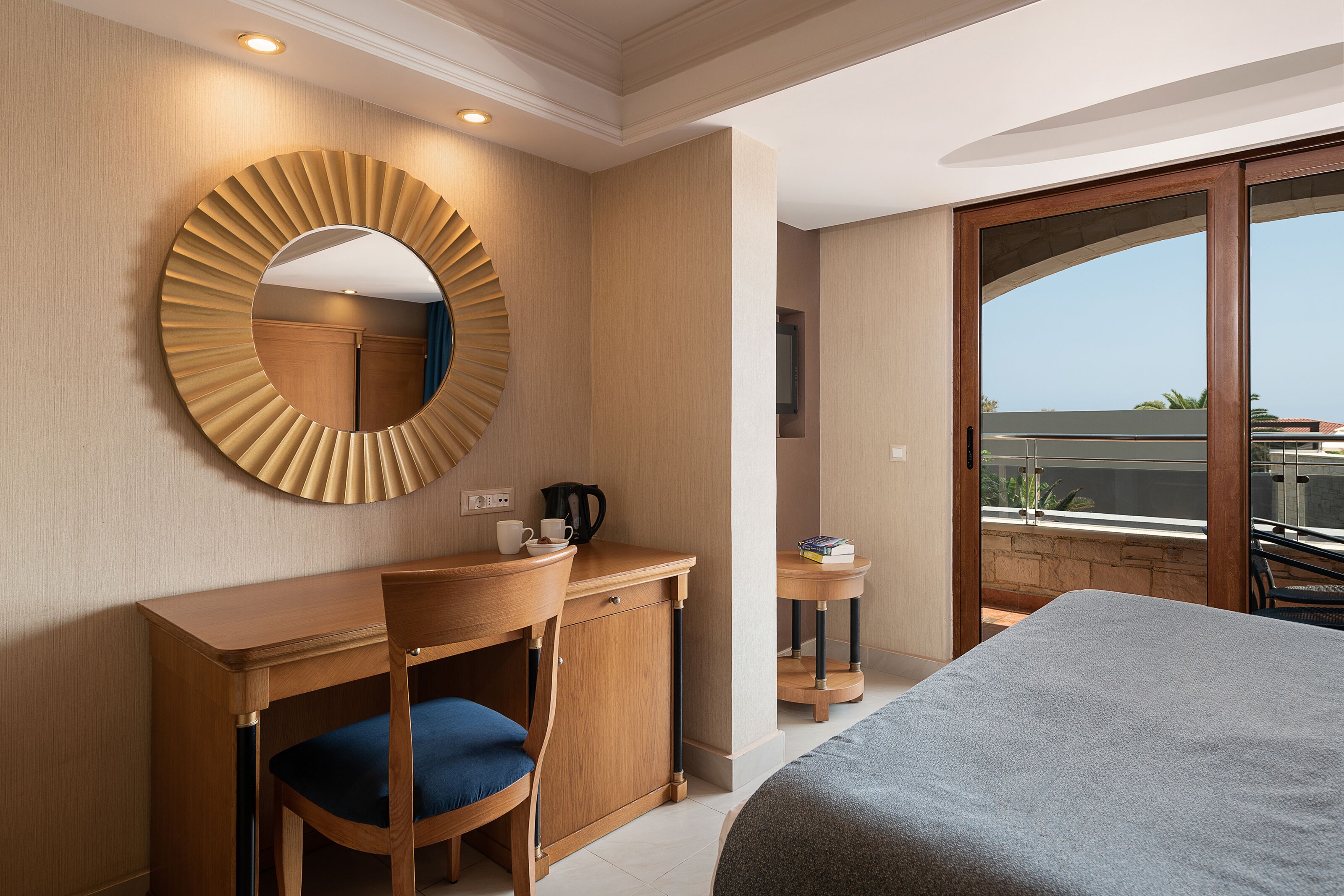 A hotel room with a wooden desk, a chair, a bed, a round mirror, and a view of the outside.