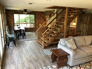 A spacious living area with a wooden staircase, a sofa, a dining table with chairs, and a large window providing natural light. - Lake front secluded house w/private boat/swim dock on 242 acre cattle ranch.  (Eagle Rock)