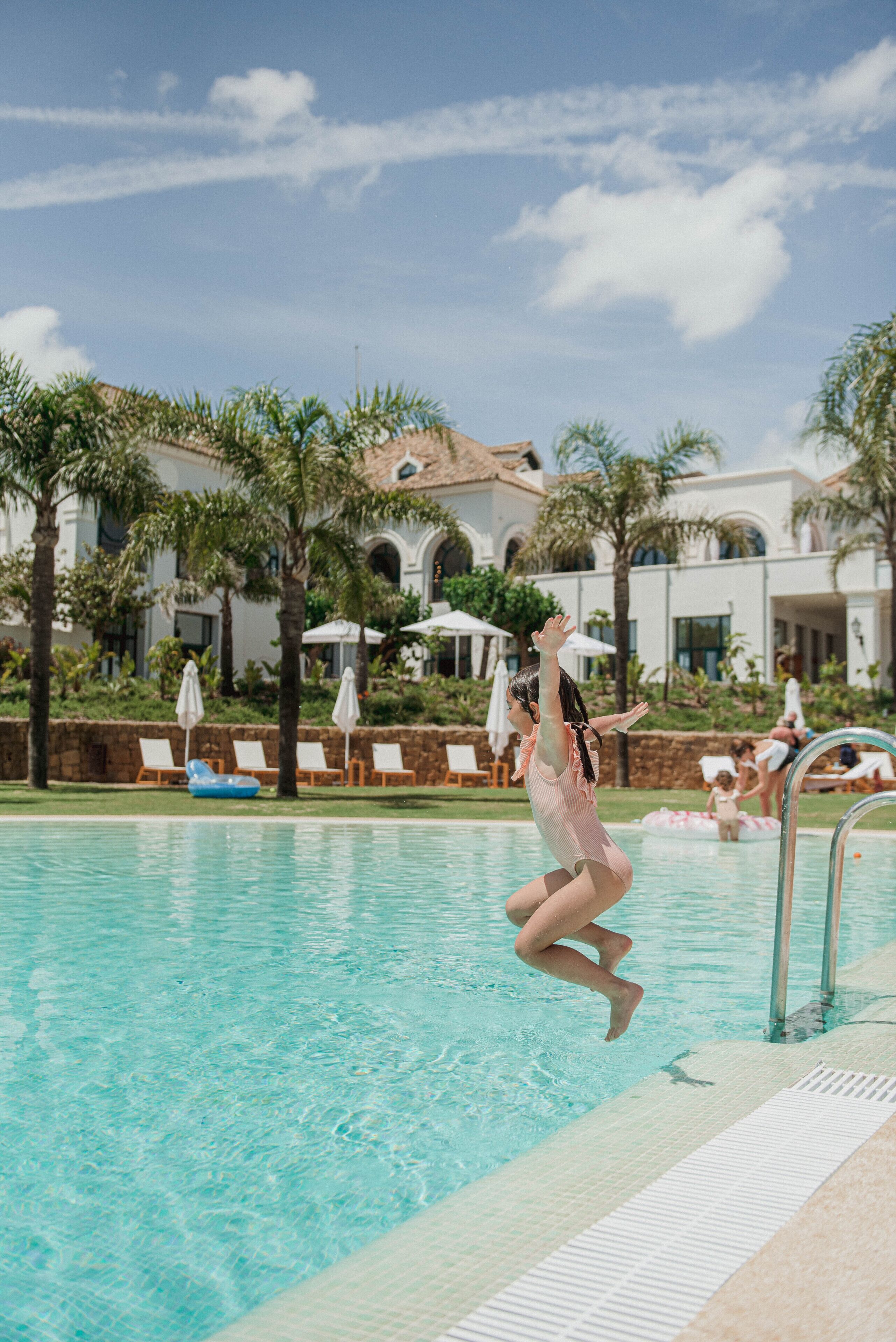 Un área de piscina con agua cristalina, sillas de descanso, sombrillas y palmeras.