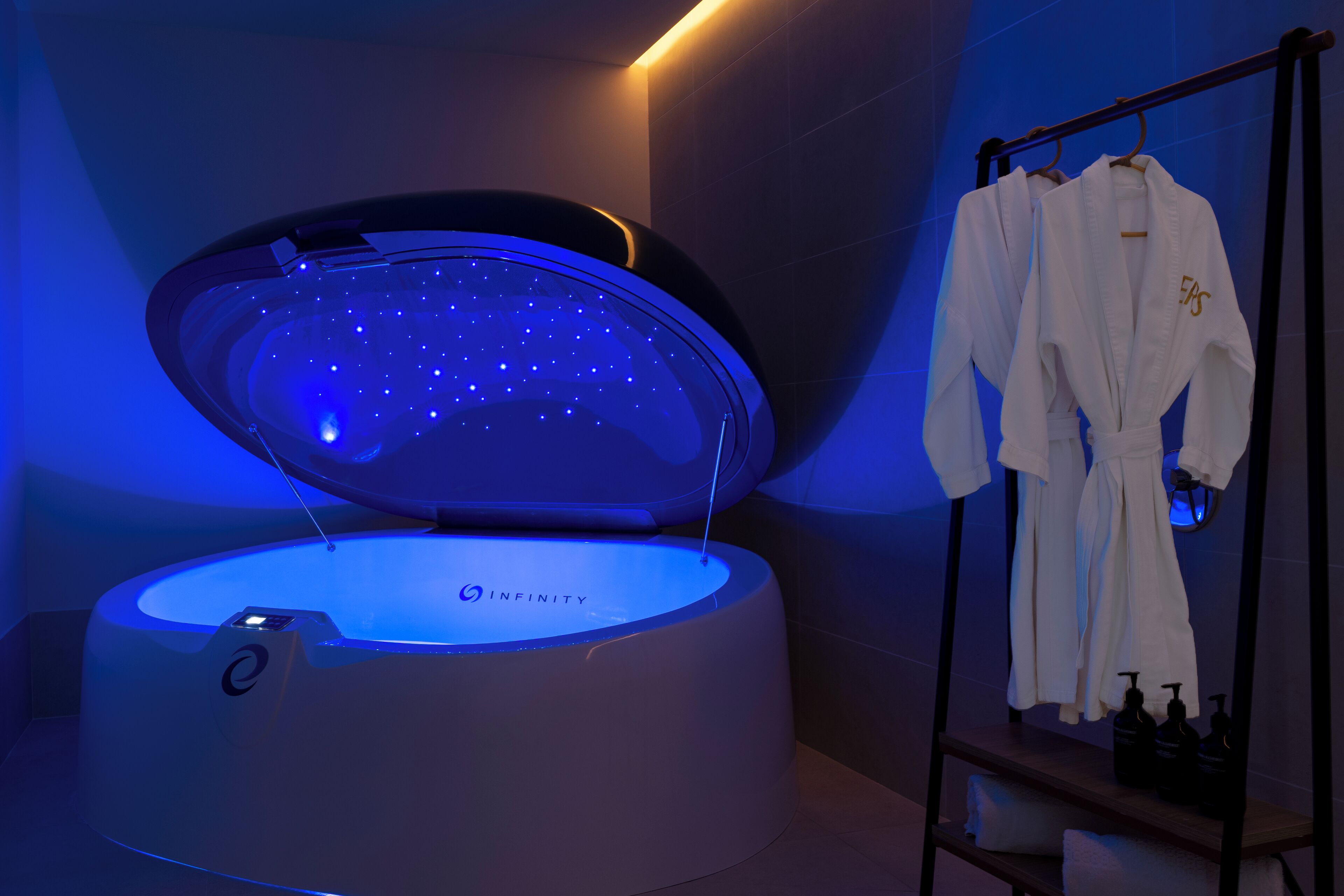 Un moderno spa con una cápsula de terapia de luz estelar, etiquetada 'INFINITY', y un perchero con batas blancas.