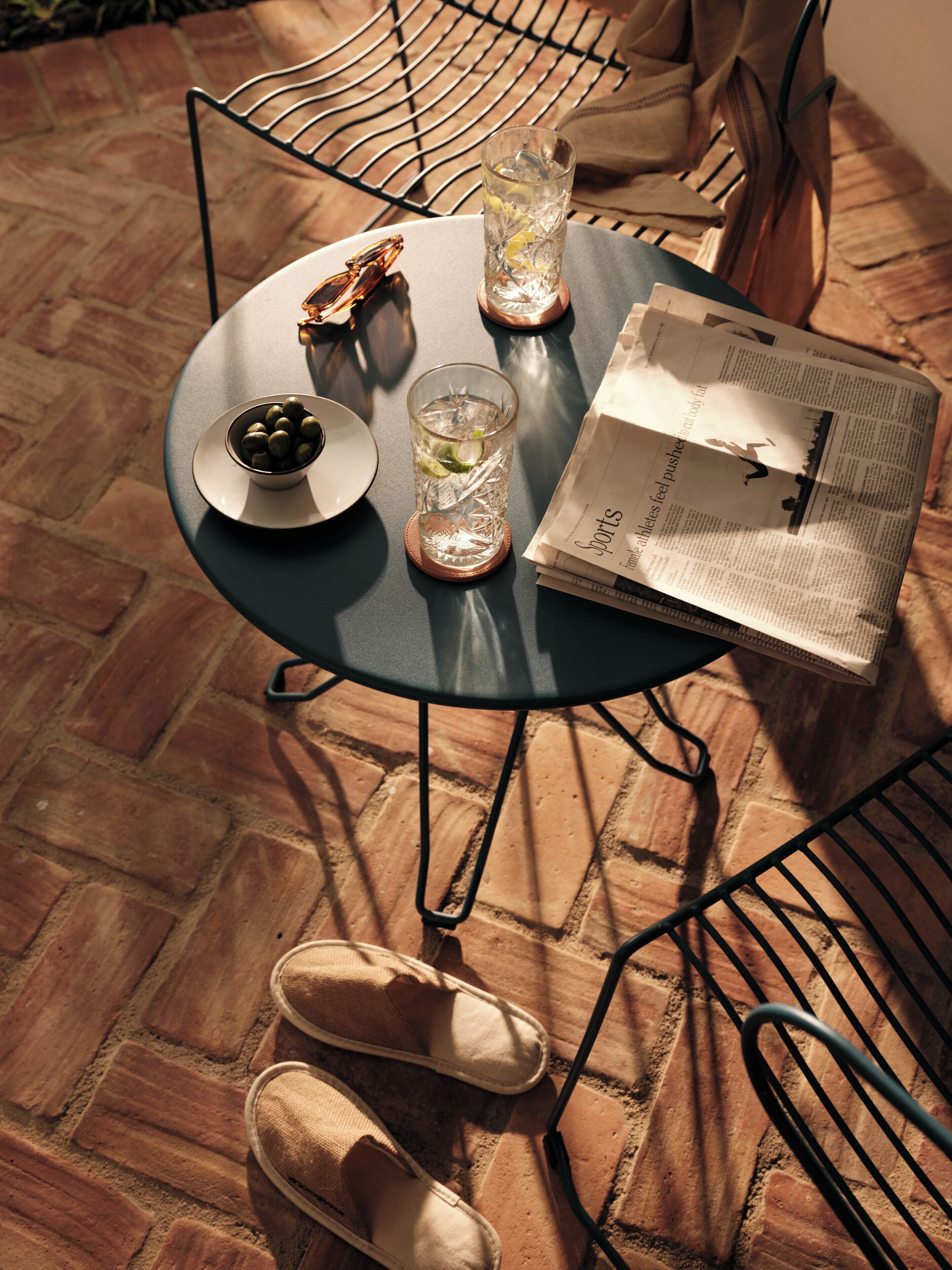 A round metal table with a glass of water, a bowl of olives, and a newspaper.