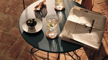 Una mesa redonda de metal con un vaso de agua, un cuenco de aceitunas y un periódico.