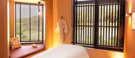Habitación de hotel con cama, ventana con persianas, banqueta con cojín y bata colgada en la pared.