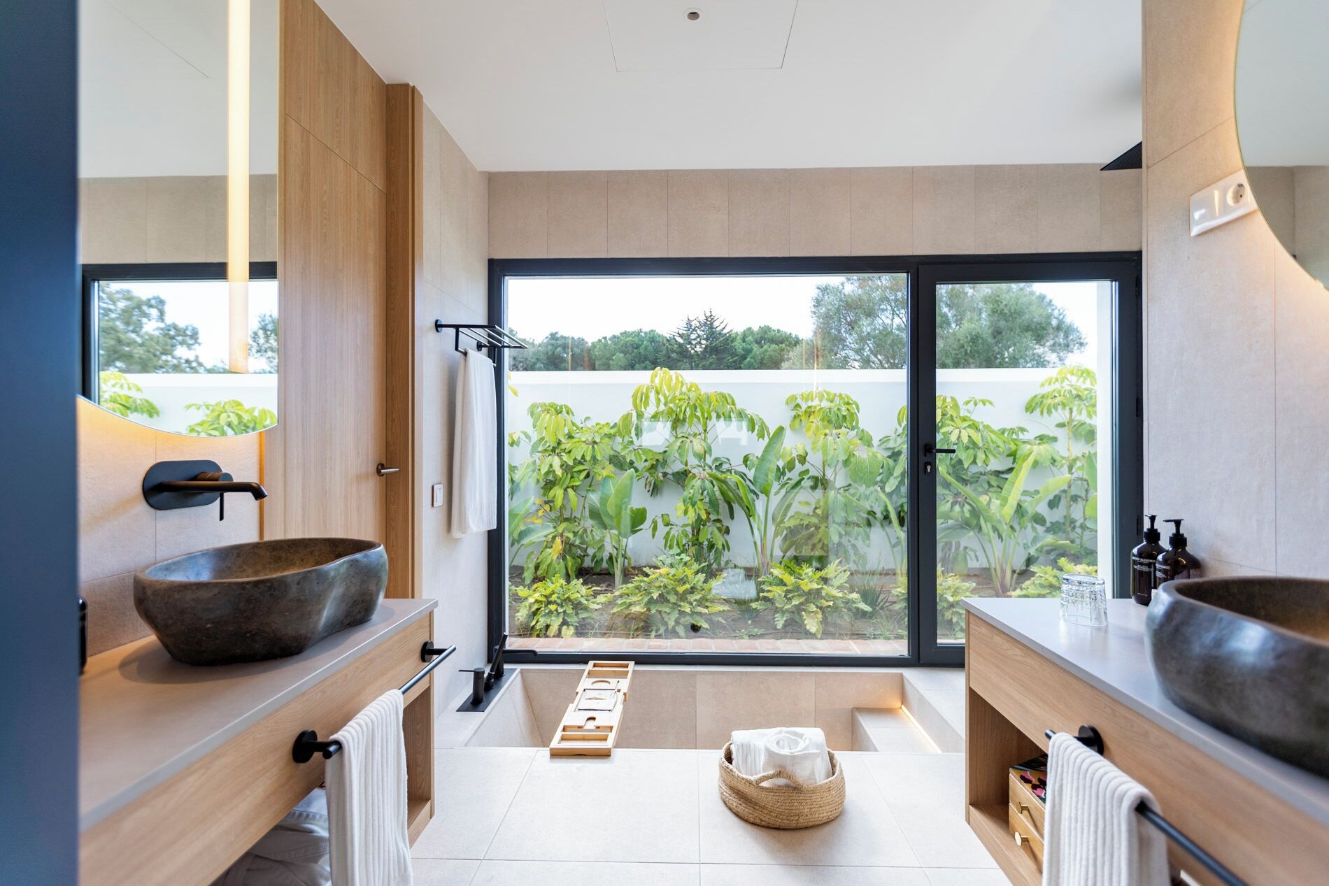 Une salle de bain moderne avec deux grands lavabos en pierre, des meubles en bois et une vue sur un jardin luxuriant, grâce à de grandes portes coulissantes.
