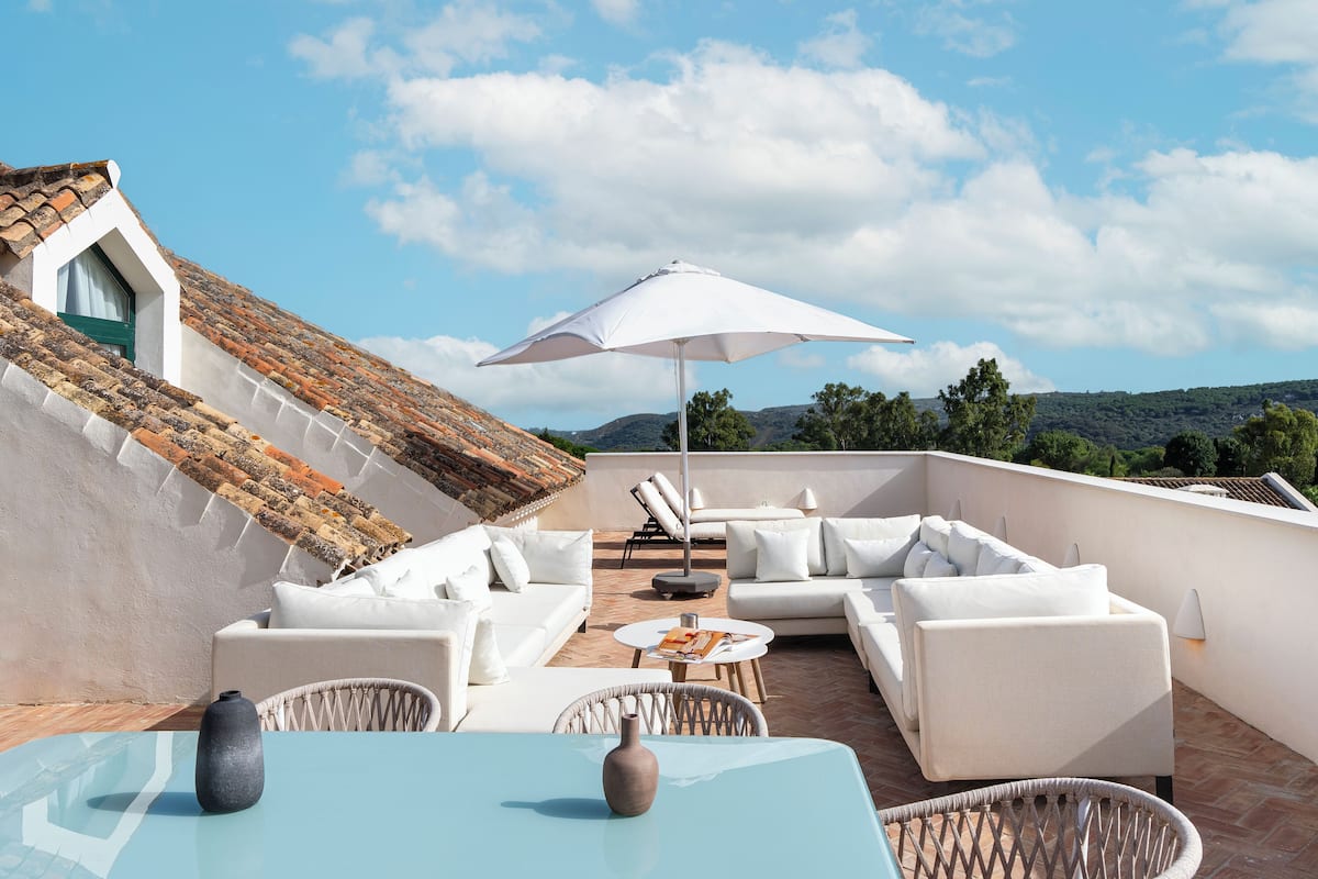 En tagterrasse med hvide møbler, blandt andet sofaer og et bord, under en stor parasol.