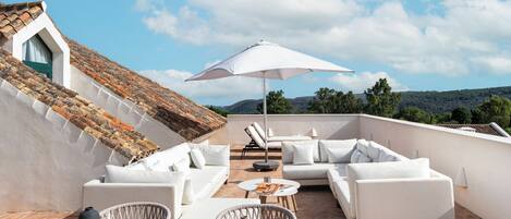 Une terrasse sur le toit, meublée de mobilier blanc, notamment de canapés et d’une table, protégée par un grand parasol.