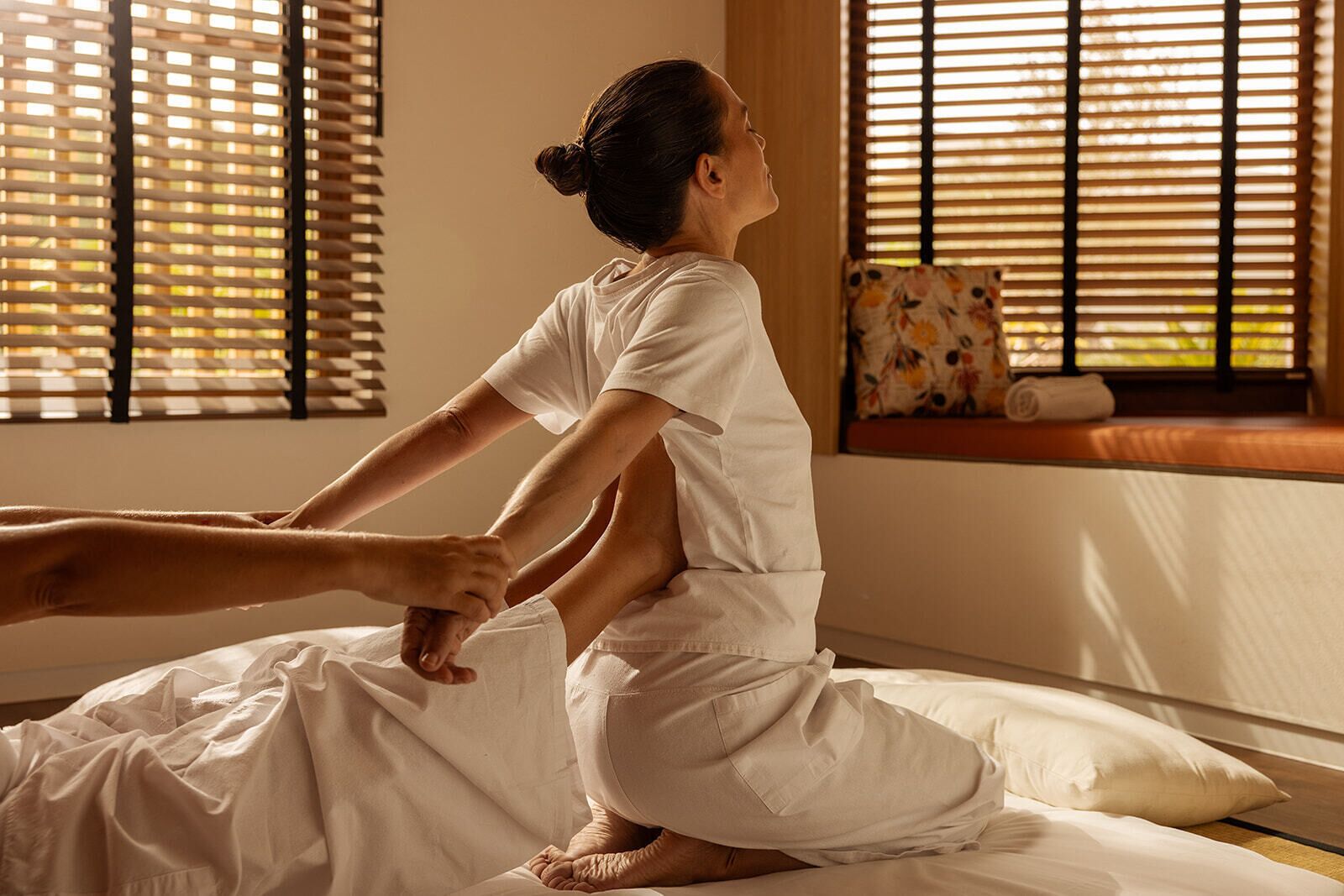 Una persona realizando un estiramiento en una cama, en una habitación con persianas de madera y una ventana con un cojín estampado con motivos florales.