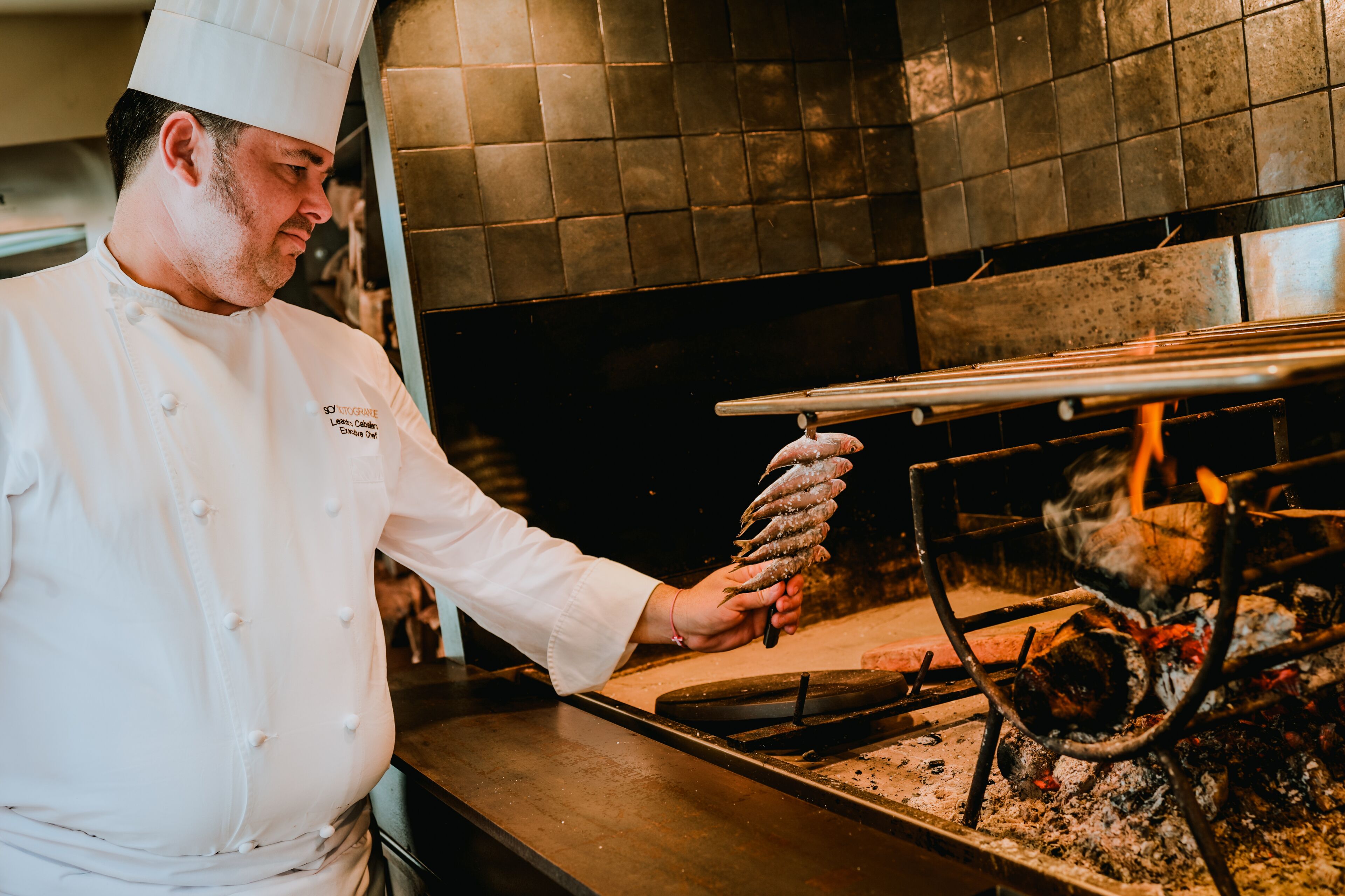 A chef in a white uniform with a tall hat, holding a tray of food over a grill with visible flames.