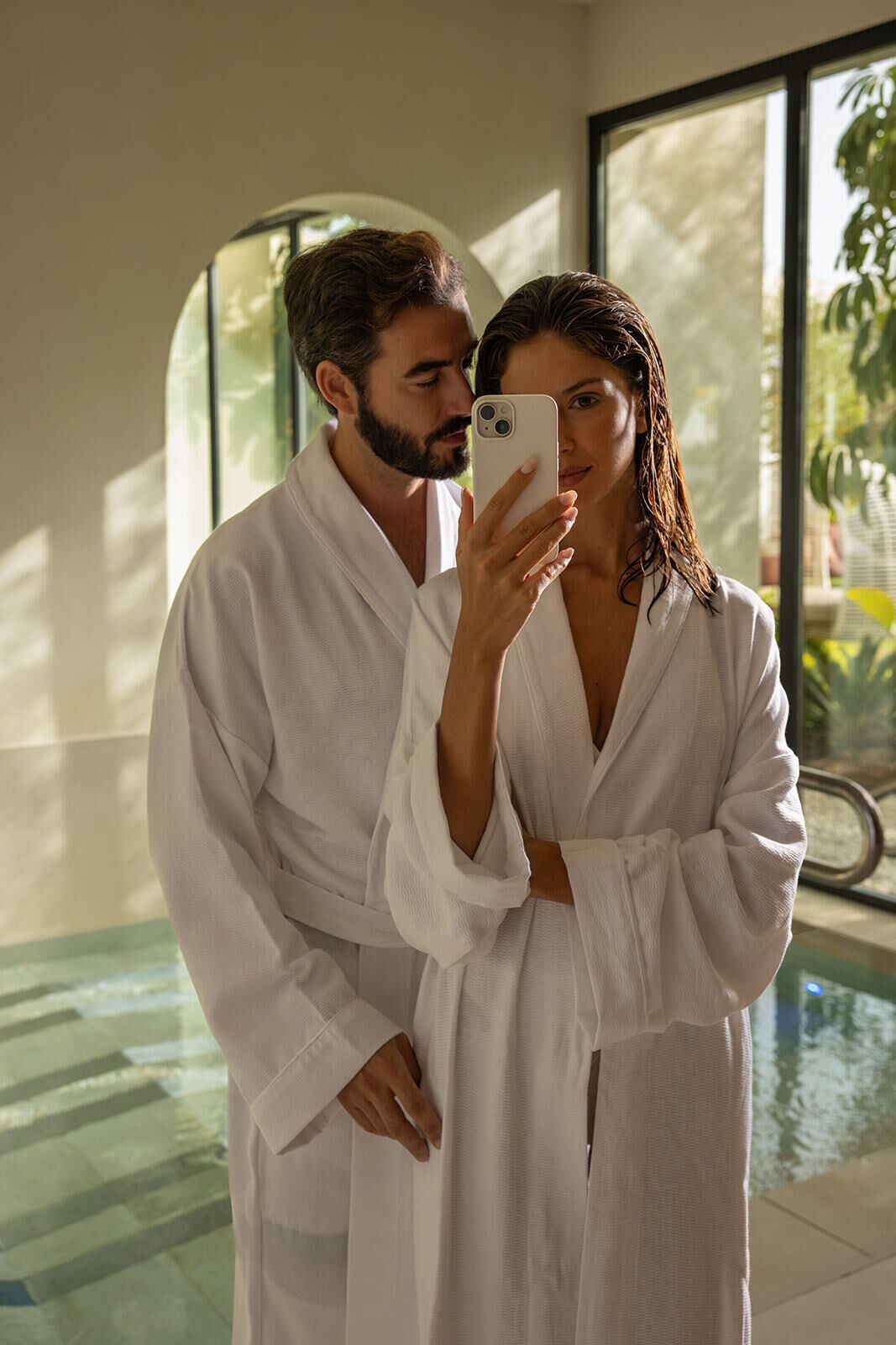 A couple in white robes by a pool with a view of greenery and a large window.