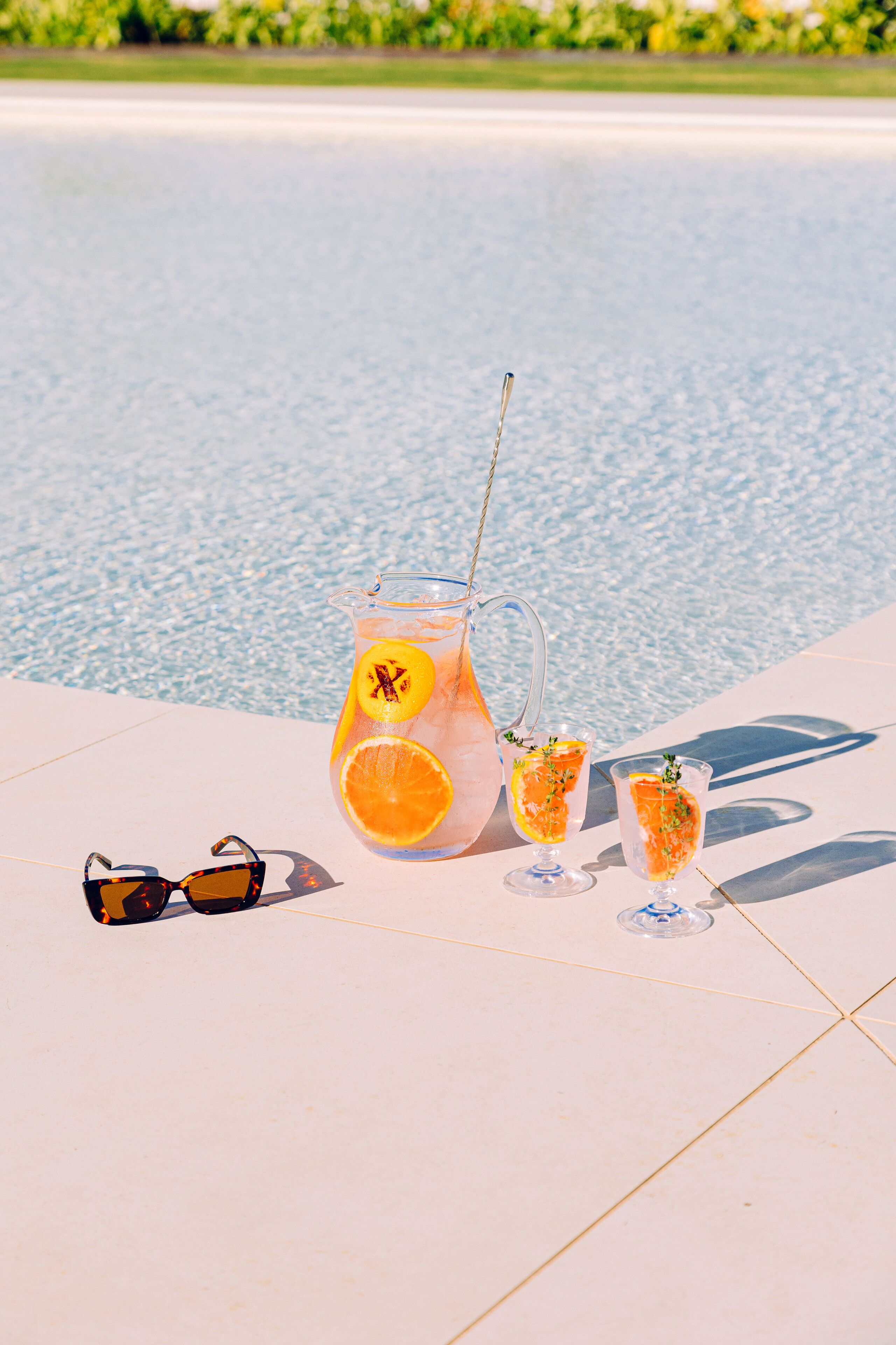 Un jarro de vidrio transparente con una pajita, dos vasos con rodajas de naranja y un par de gafas de sol sobre una superficie blanca junto a una piscina.
