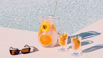 Un jarro de vidrio transparente con una pajita, dos vasos con rodajas de naranja y un par de gafas de sol sobre una superficie blanca junto a una piscina.