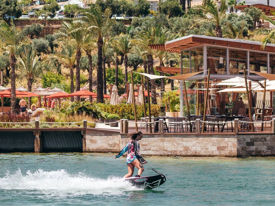 A person water skiing on a body of water with a backdrop of palm trees, a building with a glass structure, and a patio area with umbrellas.