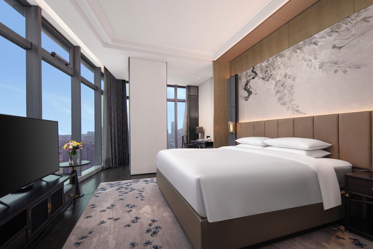 A modern hotel room with a large bed, a flat-screen TV, a desk with a chair, and a large window offering a city view.