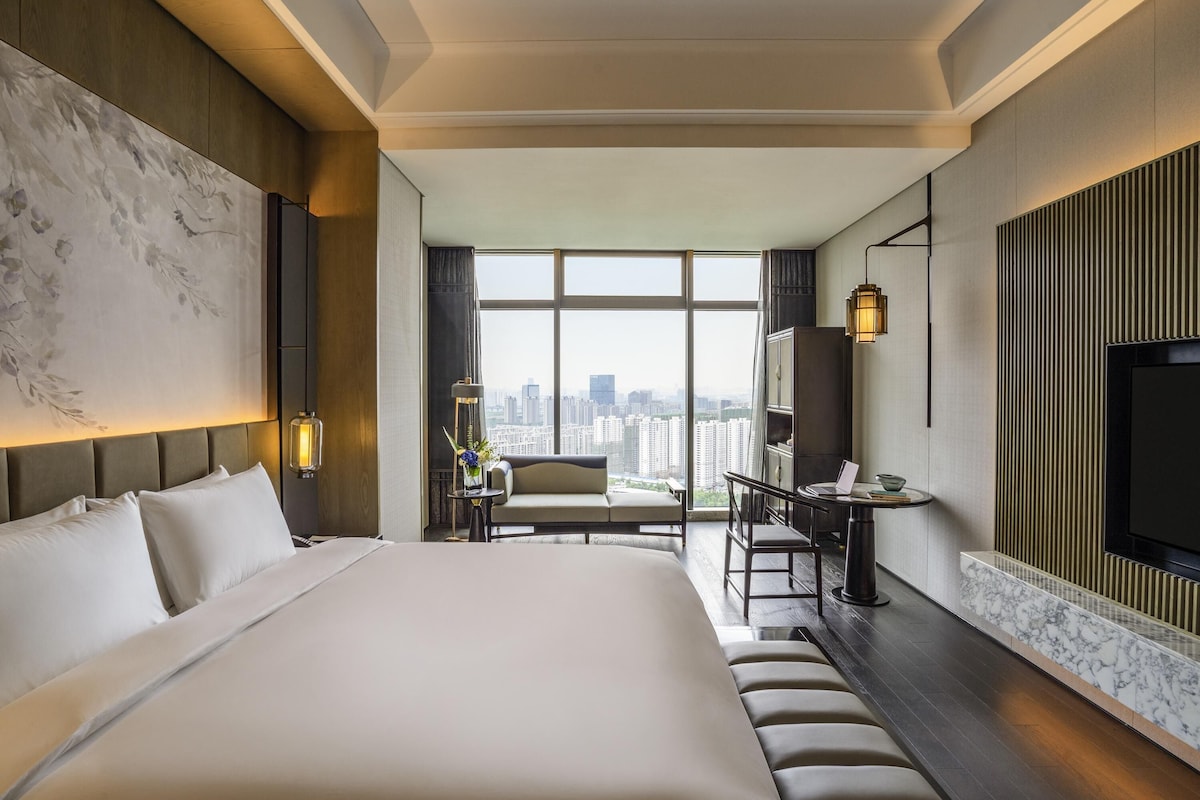 Une chambre d’hôtel moderne dotée d’un grand lit, d’un bureau avec un ordinateur portable, d’une télévision et offrant une vue sur la ville grâce à de grandes fenêtres.