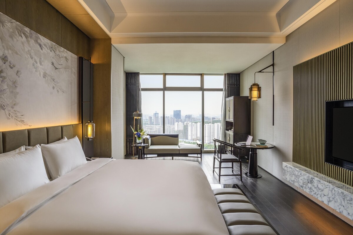 Een moderne hotelkamer met een groot bed, een bureau met een laptop, een televisie en uitzicht op de stad door grote ramen.