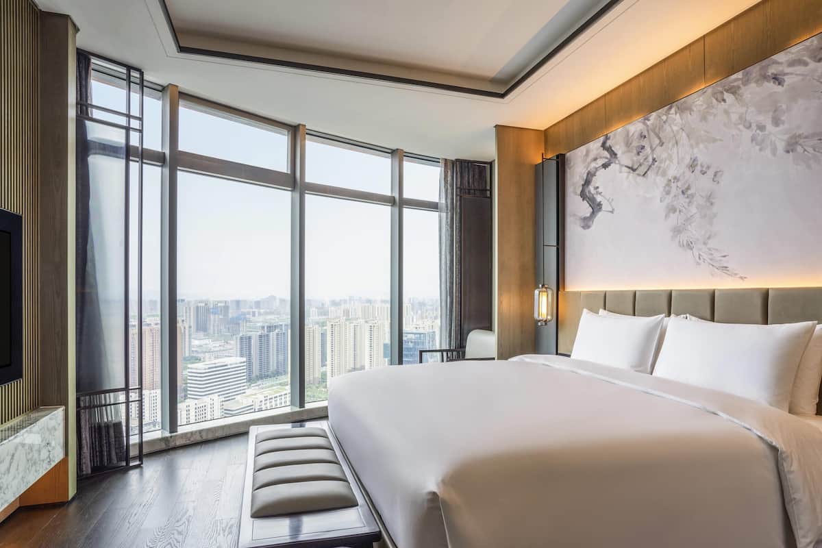 Une chambre d’hôtel moderne dotée d’un grand lit, d’une banquette, d’un téléviseur et offrant une vue sur la ville grâce à de grandes fenêtres.