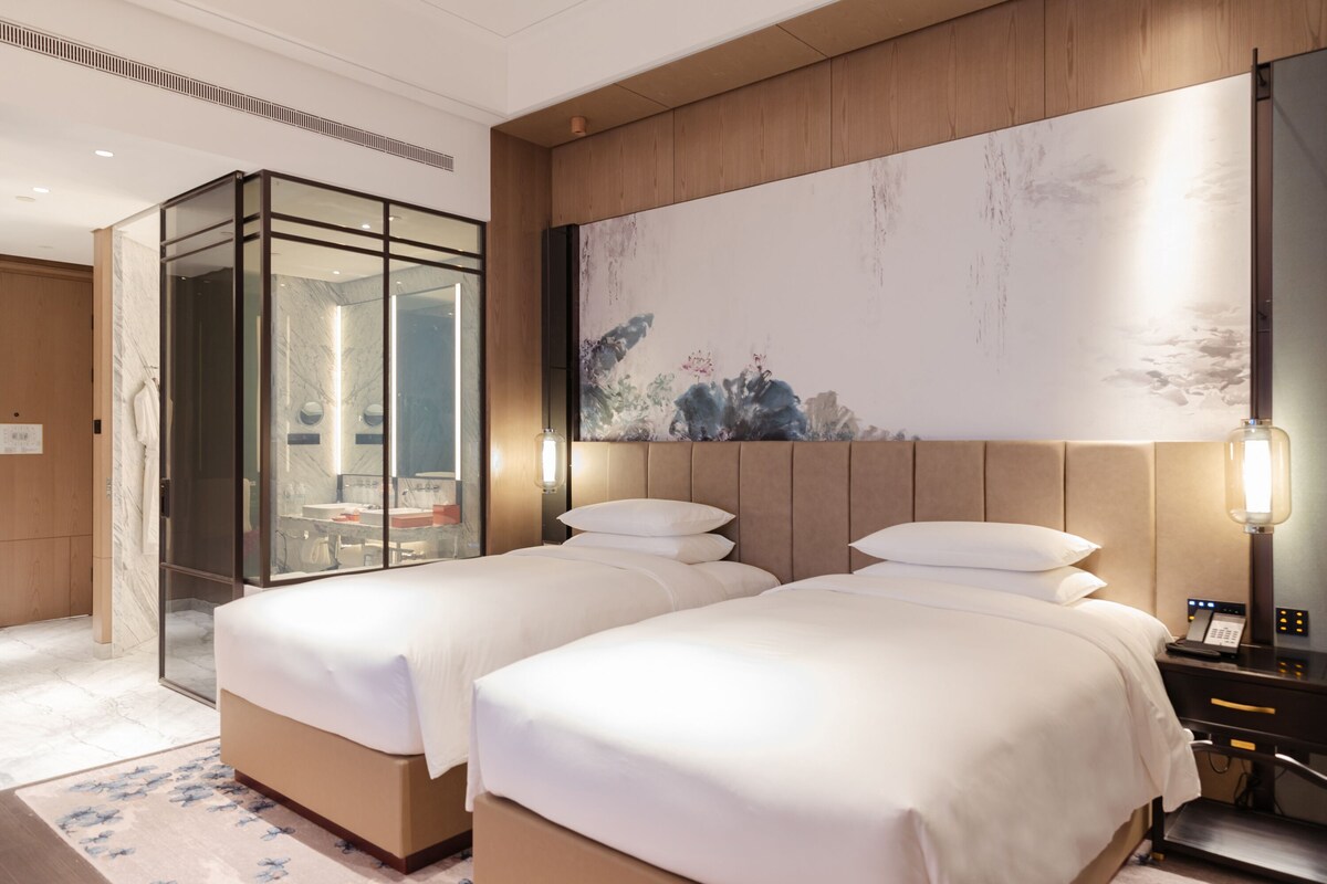 A hotel room with two beds, a glass-enclosed shower, a large abstract painting, and a bedside table with a lamp.