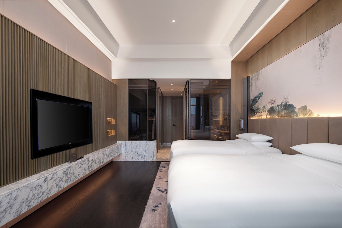 A modern hotel room with a large flat-screen TV, a marble accent wall, a large bed with white linens, and a view of the cityscape through the window.