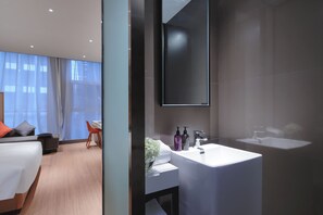 Quarto de hotel moderno com uma cama, escrivaninha, cadeira, banheiro com pia e espelho, e uma janela grande.