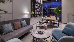 A modern living room with a sofa, coffee table, and dining area. There is a bookshelf and a large window with a city view.