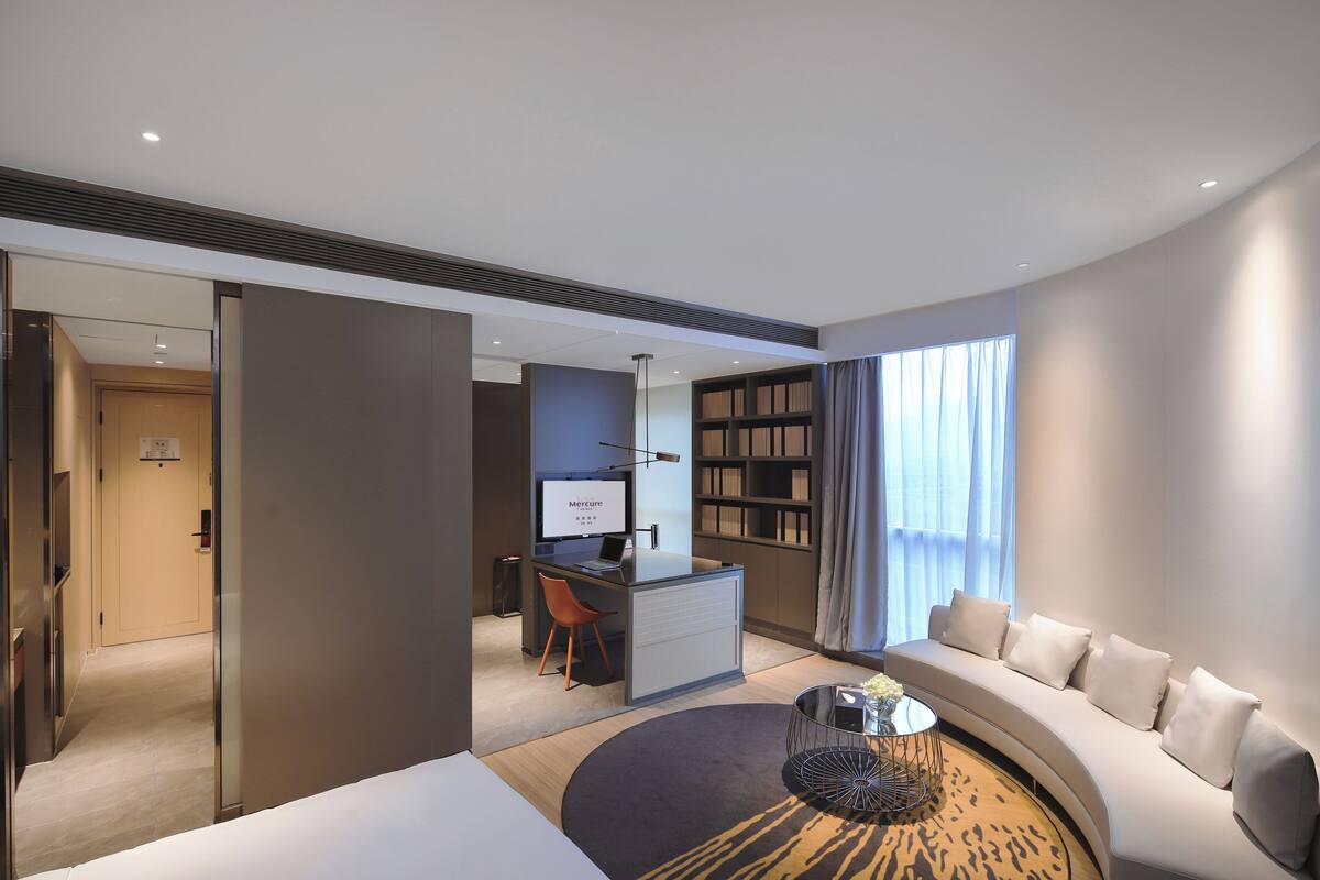 Una habitación de hotel moderna con un sofá curvo, un escritorio pequeño con una computadora, una estantería y un amplio ventanal con cortinas.