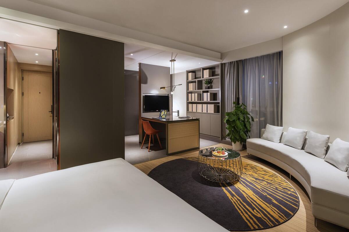 Una habitación de hotel moderna con un sofá curvo, un comedor con mesa redonda, un televisor y una estantería.