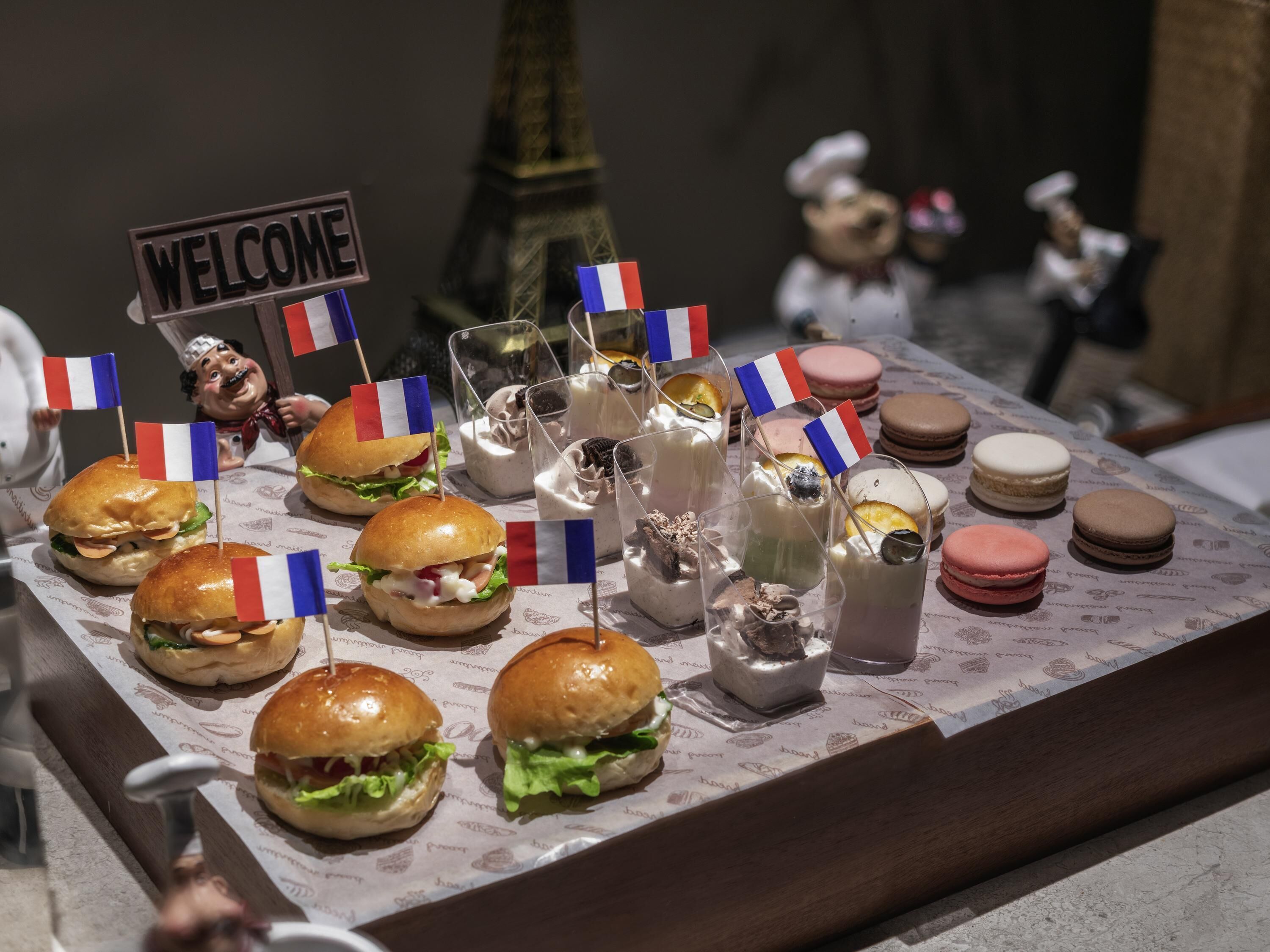 Una mesa con diversos alimentos, entre ellos sándwiches, macarons y postres, cada uno decorado con una pequeña bandera francesa.