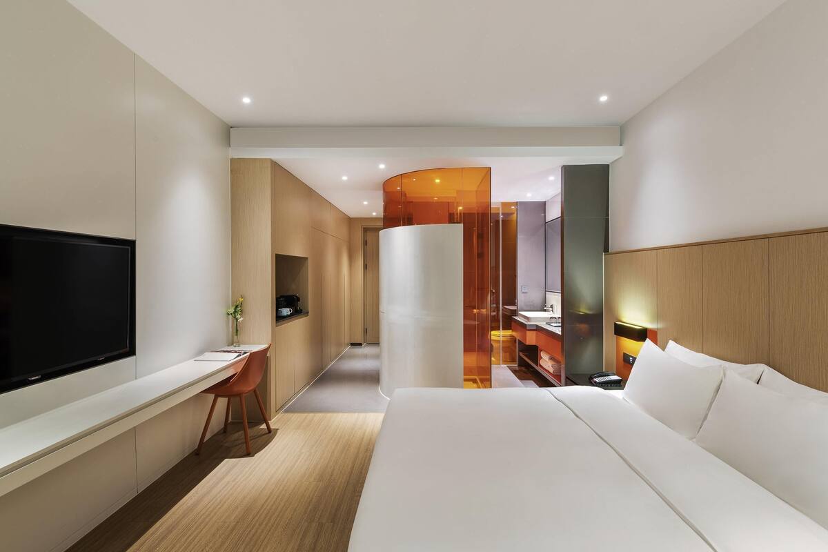 Habitación de hotel moderna con una cama grande, un escritorio con silla, un televisor y un baño visible a través de un panel de vidrio.