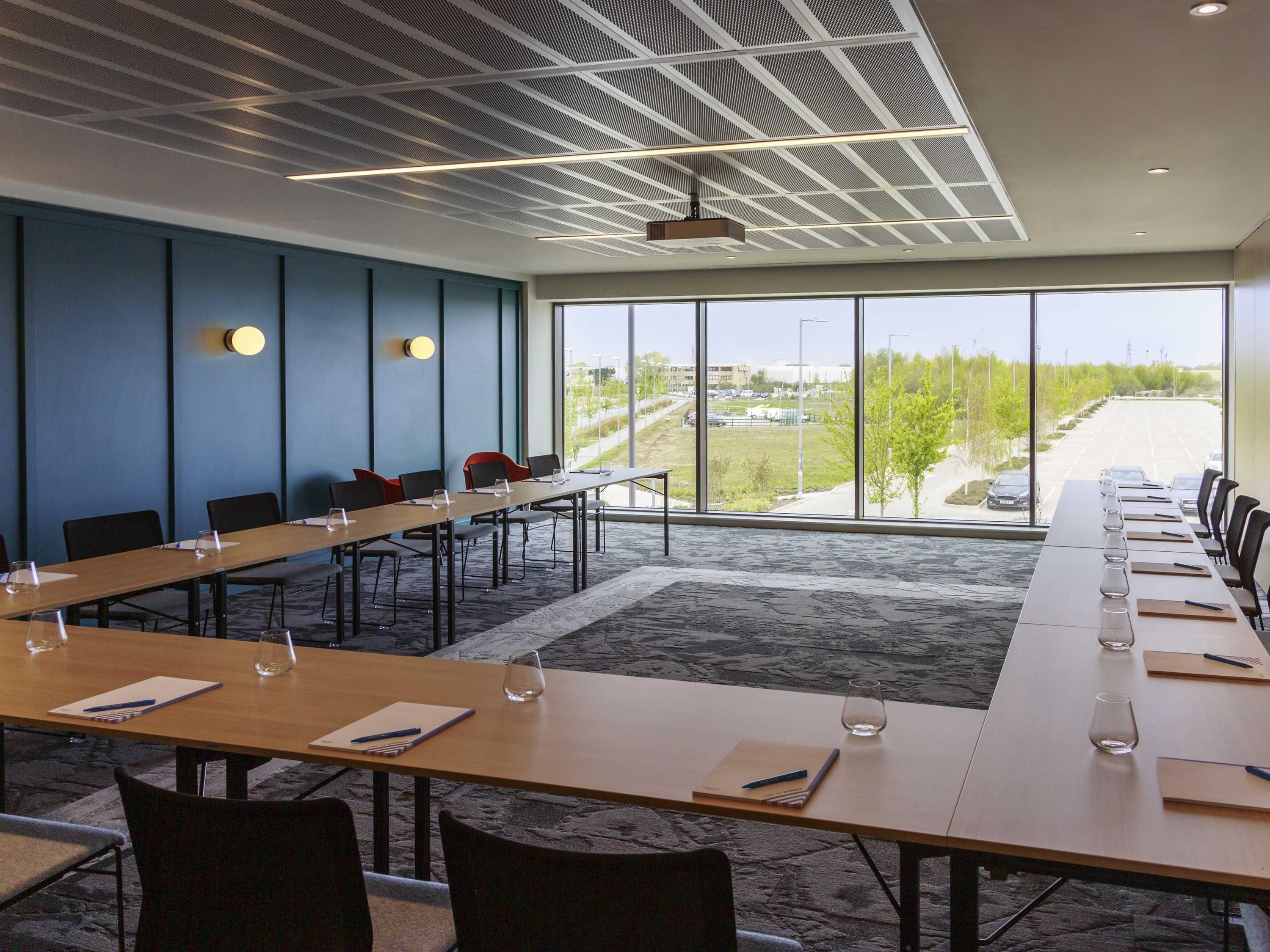 Un salón de conferencias con una larga mesa, sillas y un amplio ventanal que ofrece vistas a un estacionamiento y zonas verdes.