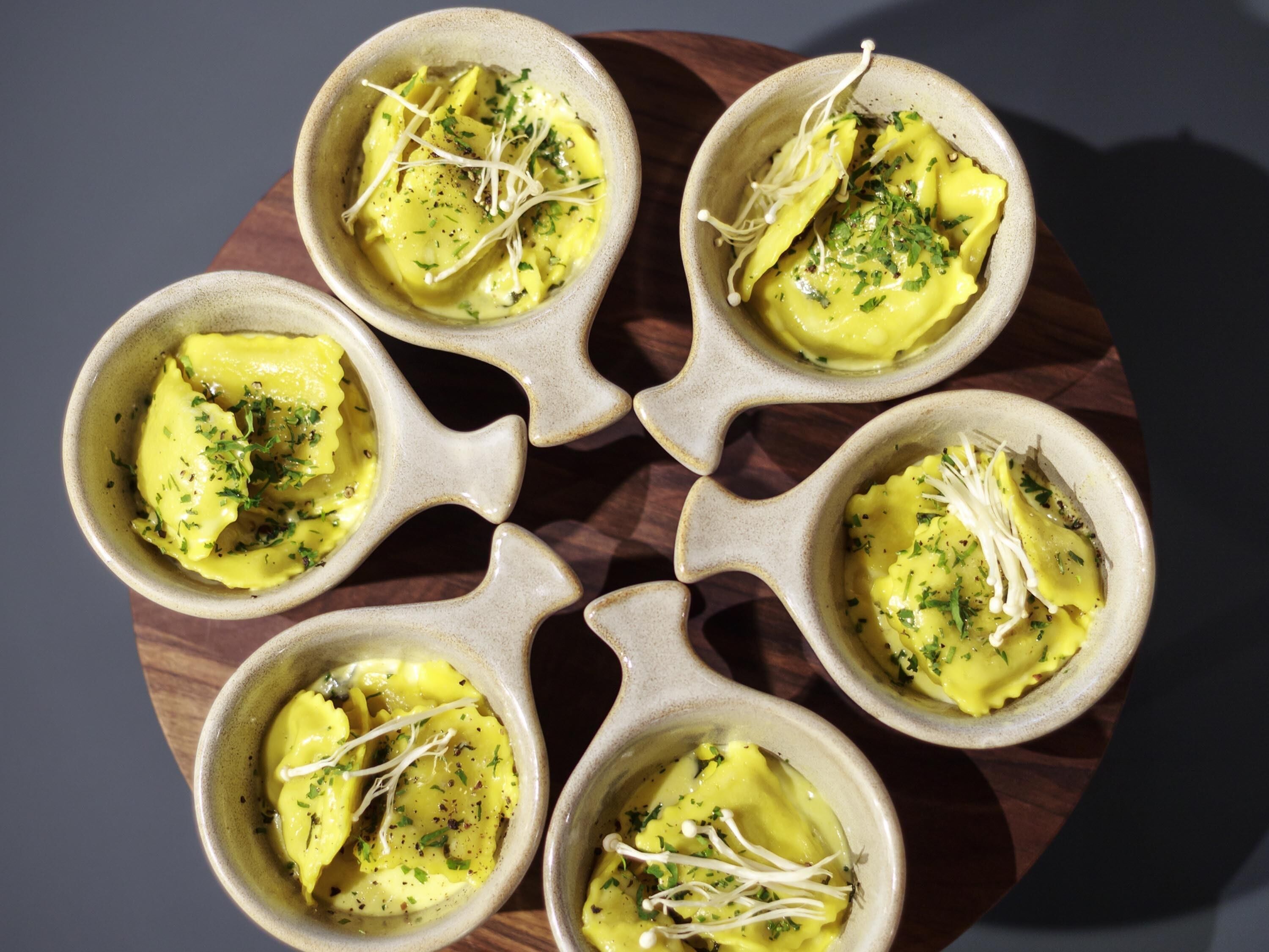 Six bowls of pasta with a creamy sauce, garnished with herbs and sprouts, arranged on a wooden board.
