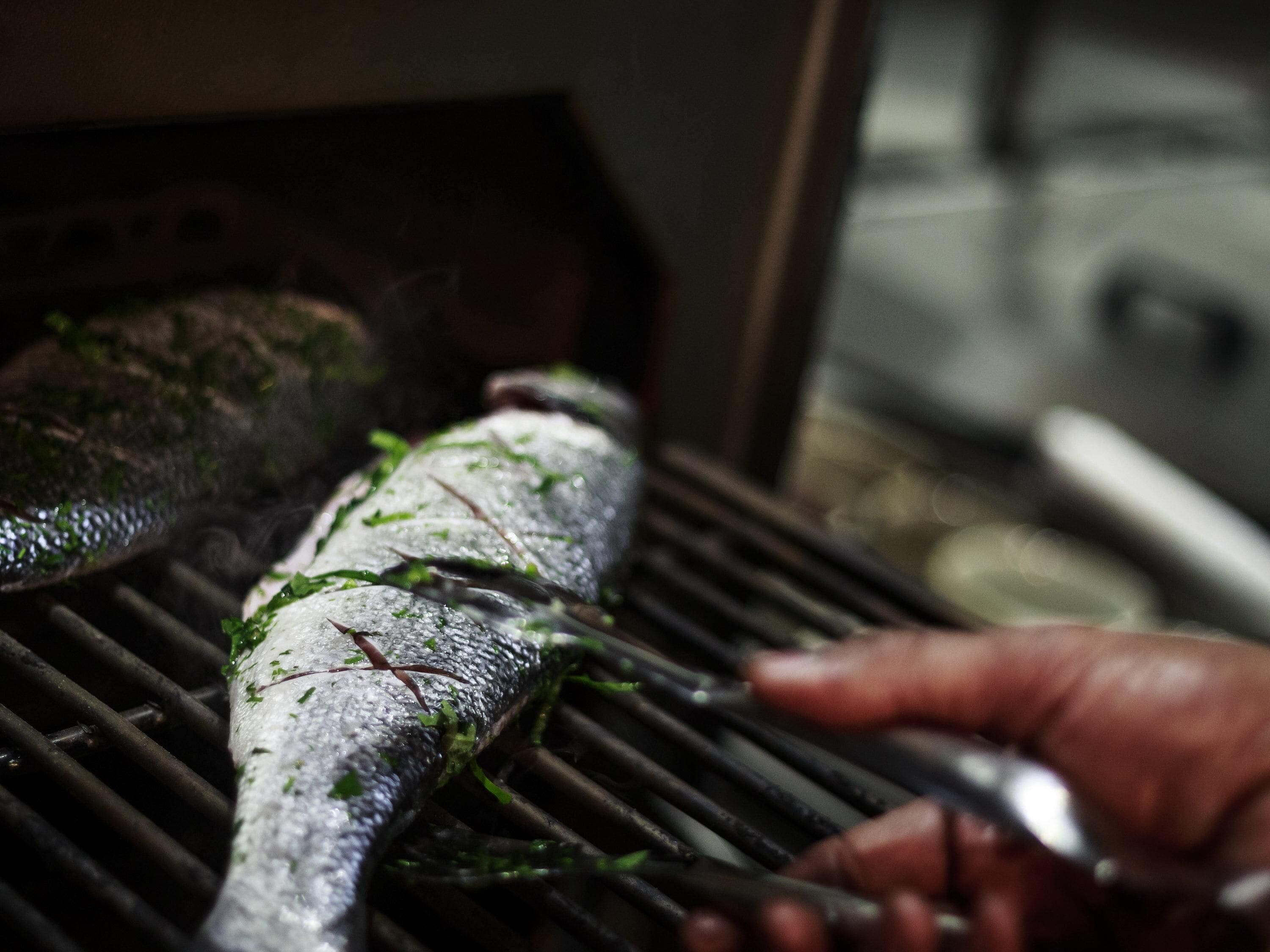 A fish being grilled on a grill with a hand holding tongs.