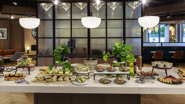 A buffet setup with various food items, a potted plant, and modern lighting fixtures. - Novotel Cambridge North (Cambridge)