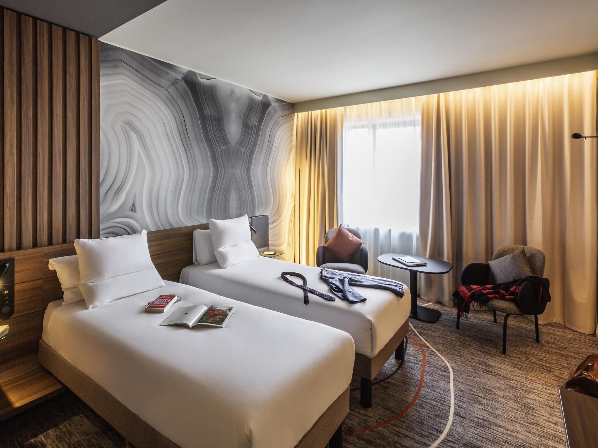 A hotel room with two beds, a small table, a chair, and a large window with curtains.