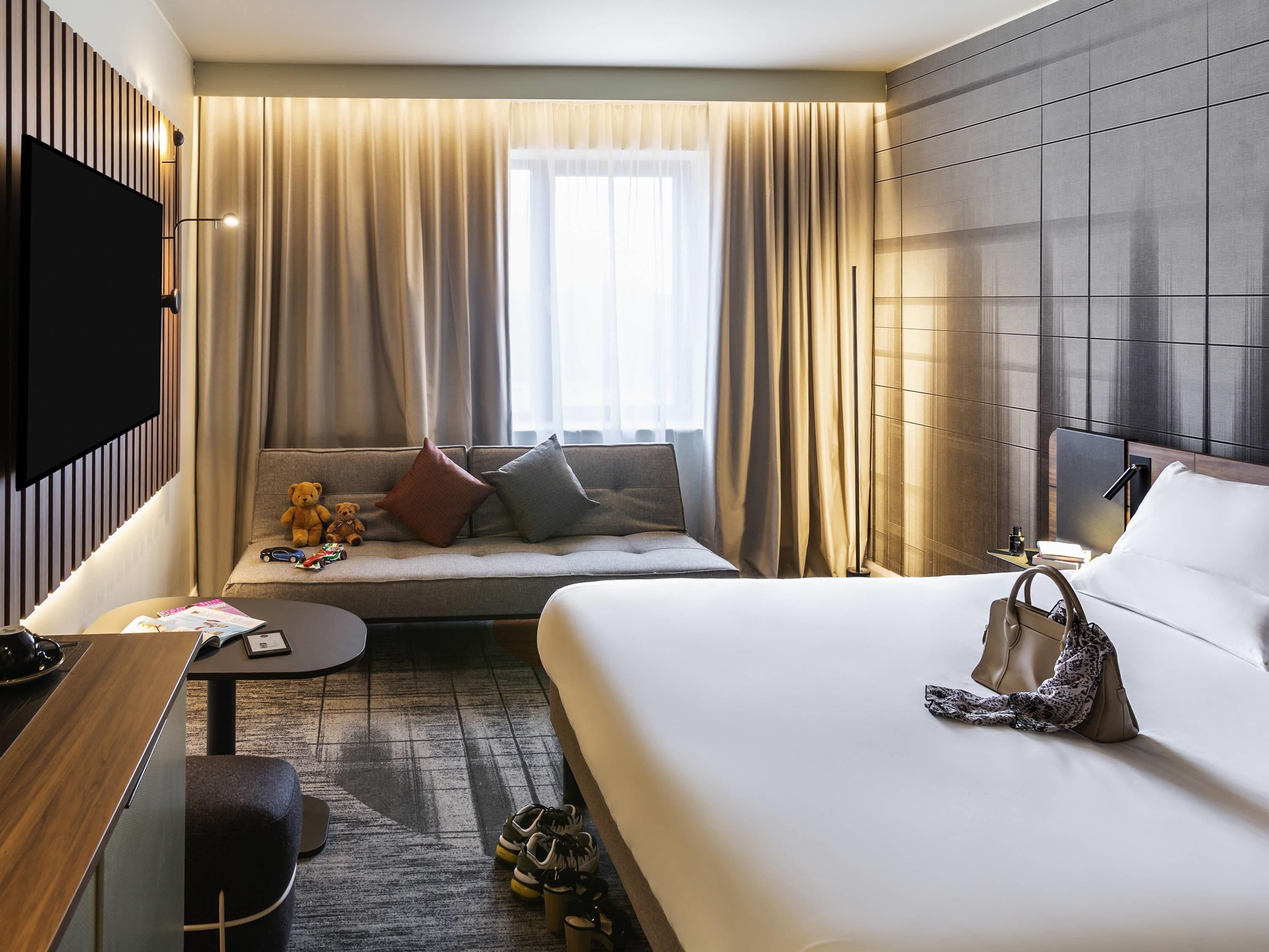 A hotel room with a bed, a sofa with pillows, a TV, a small table, and a pair of shoes on the floor.