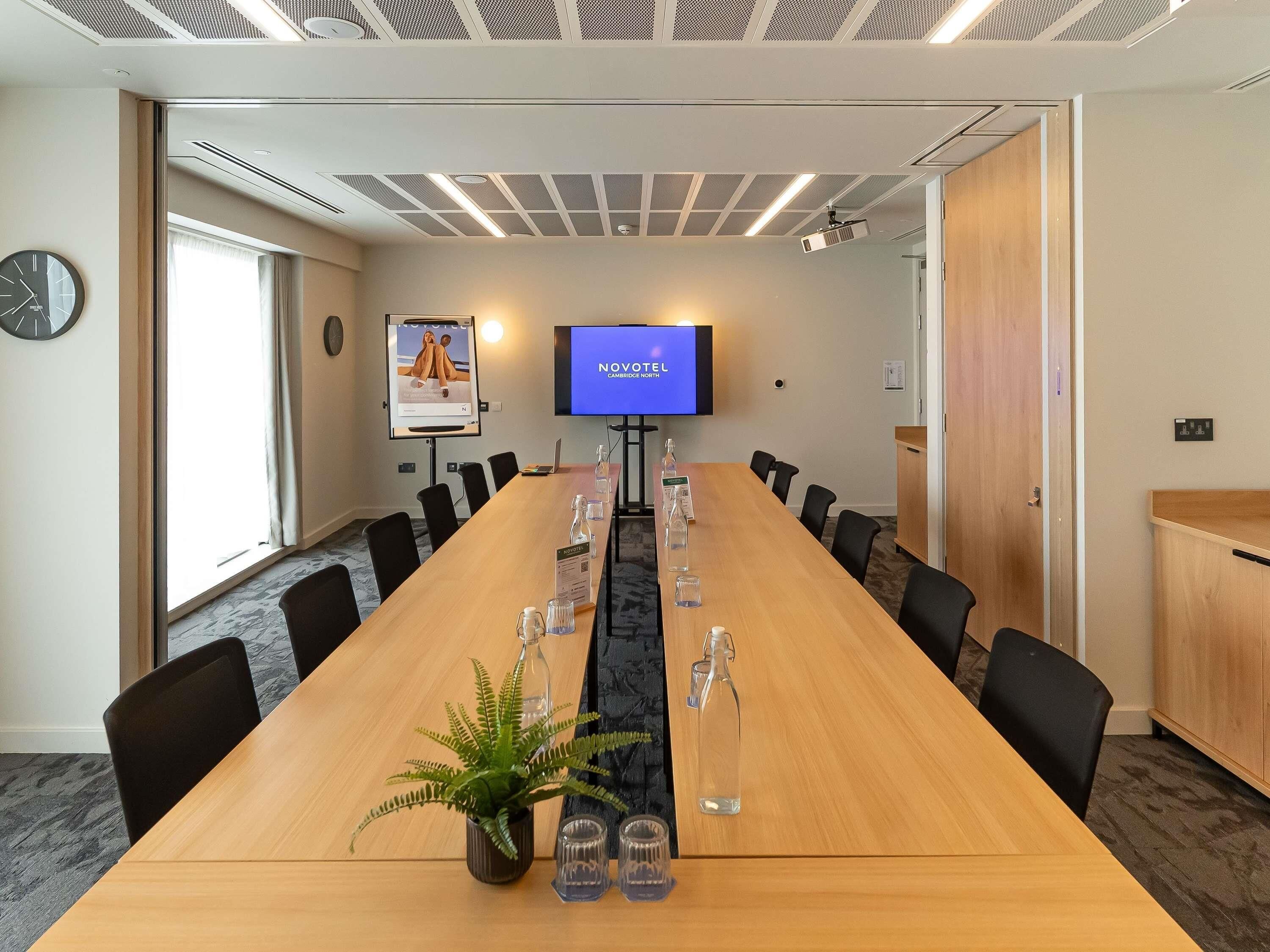 Une salle de conférence avec une longue table, des chaises, un écran de télévision, un projecteur et une horloge au mur.
