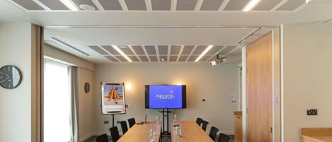 Un salón de conferencias con una larga mesa, sillas, una pantalla de televisión, un proyector y un reloj en la pared.