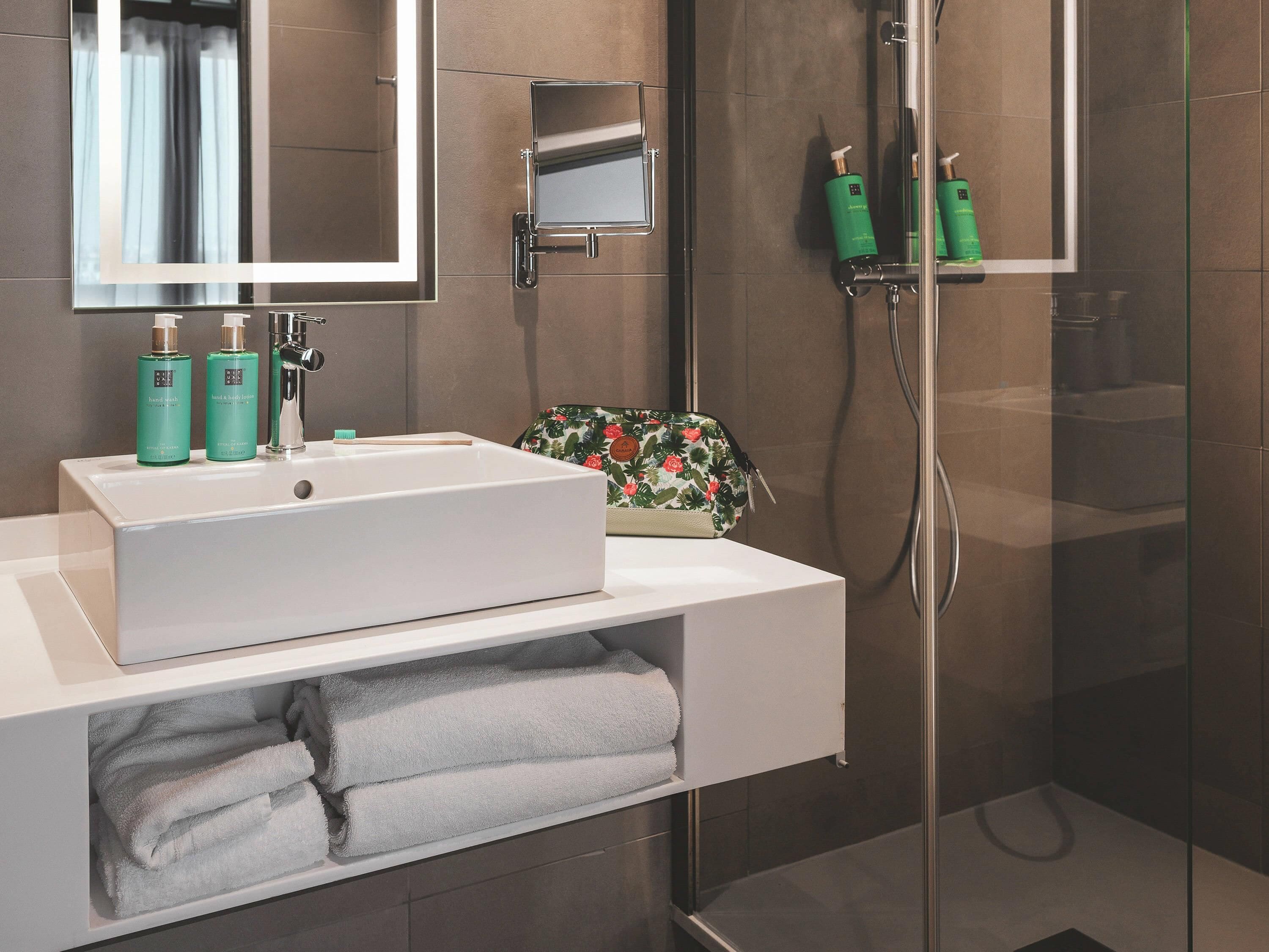 A modern bathroom with a white sink, mirror, and shower. There are toiletries on the sink and a towel rack with towels.