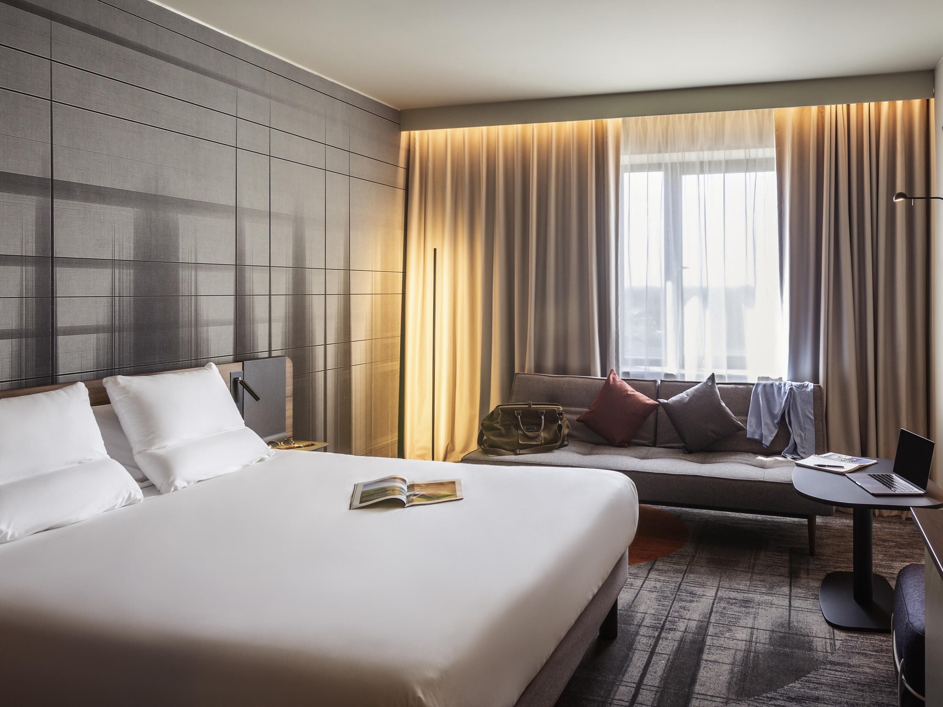 Quarto de hotel com uma cama grande, um sofá, uma escrivaninha com um laptop e uma janela com cortinas.