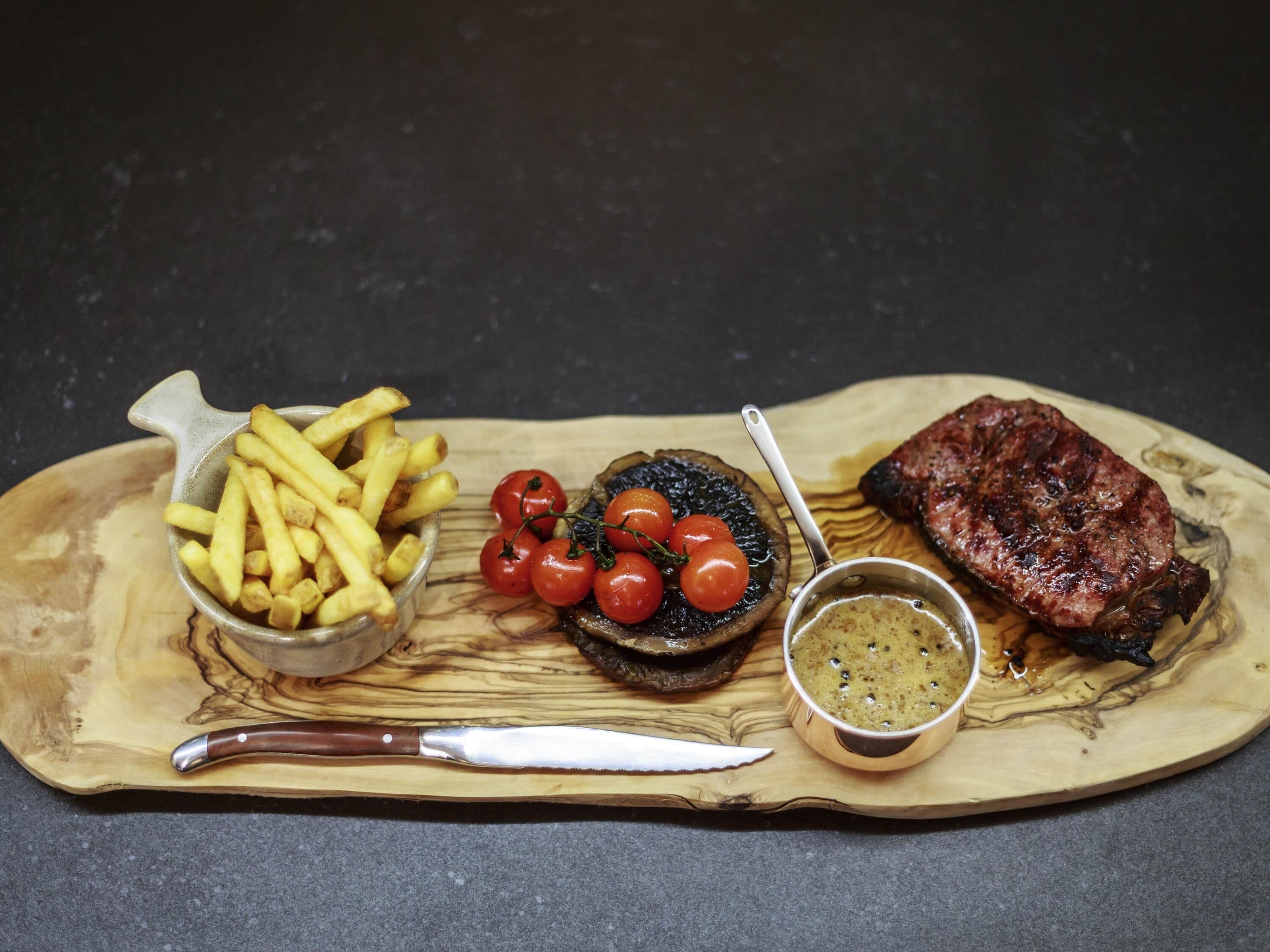 Une planche de service en bois sur laquelle sont disposés un steak, des frites, des tomates cerises et une sauce pour tremper.