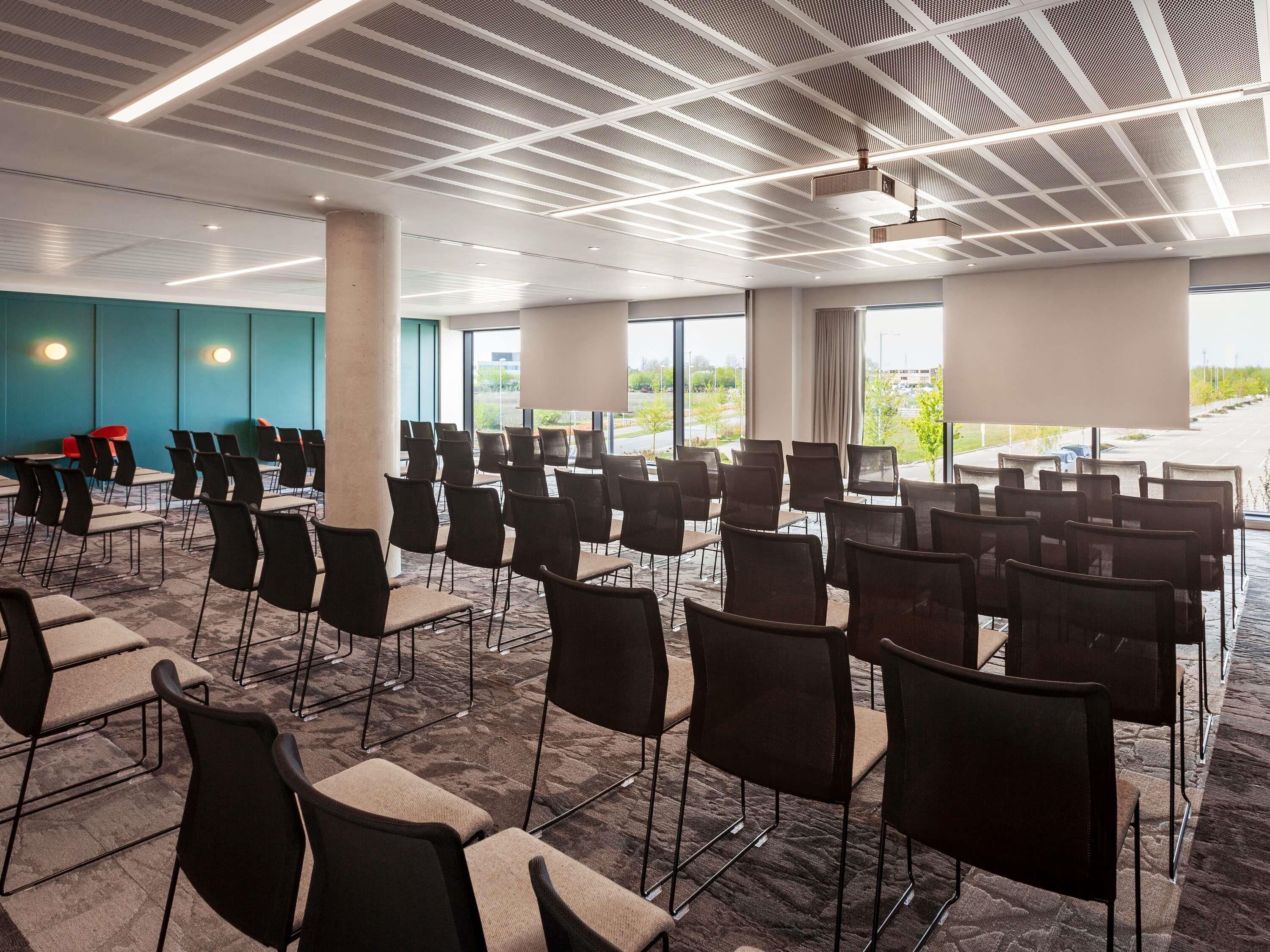 A conference room with rows of chairs, a modern ceiling design, and large windows offering a view of the outdoors.